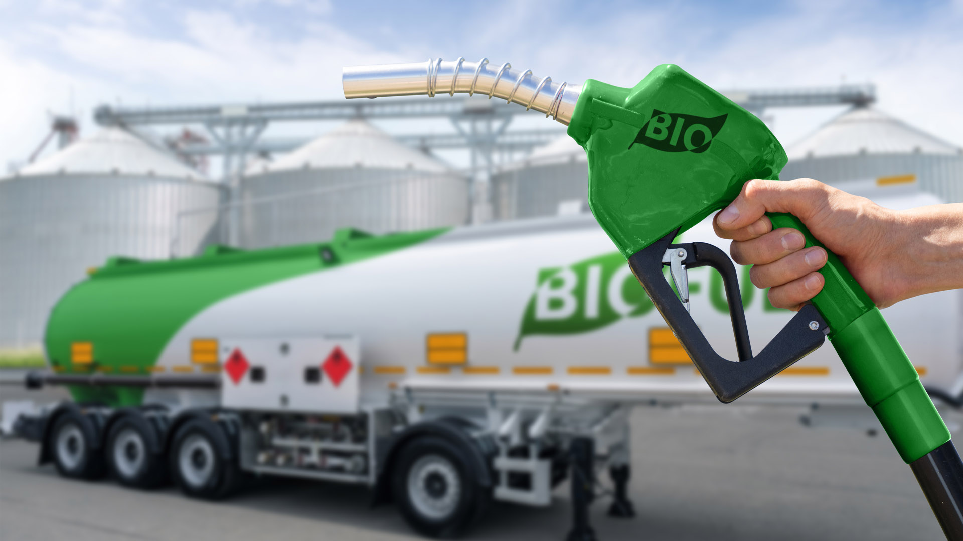 Photograph showing a close-up of a hand holding a green fuel pump nozzle labeled "BIO" in front of a large tanker truck with "BIO" written on its side. The image highlights biofuel transportation and usage, emphasizing eco-friendly fuel alternatives.