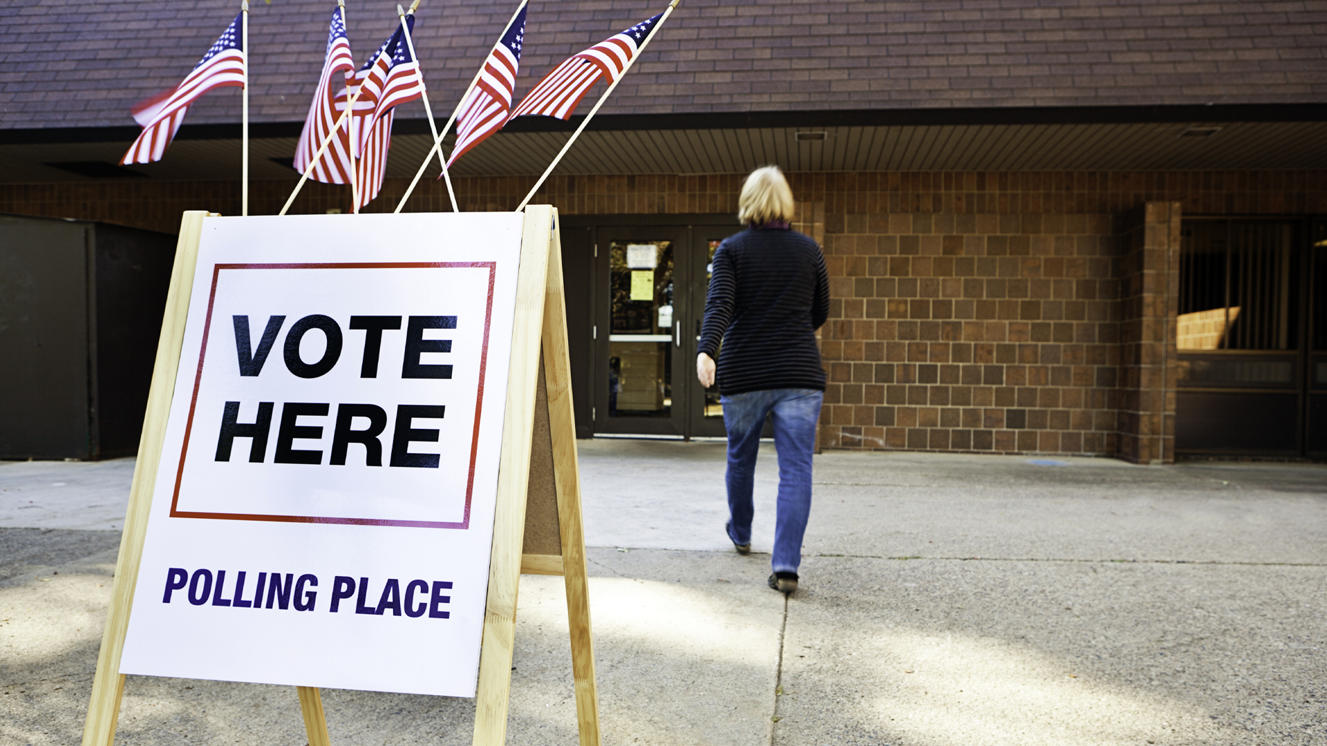 An outdoor polling place sign beside a brick building. White sign reads “VOTE HERE” and “POLLING PLACE,” with red-and-blue borders and several American flags above; a person walks toward entrance.