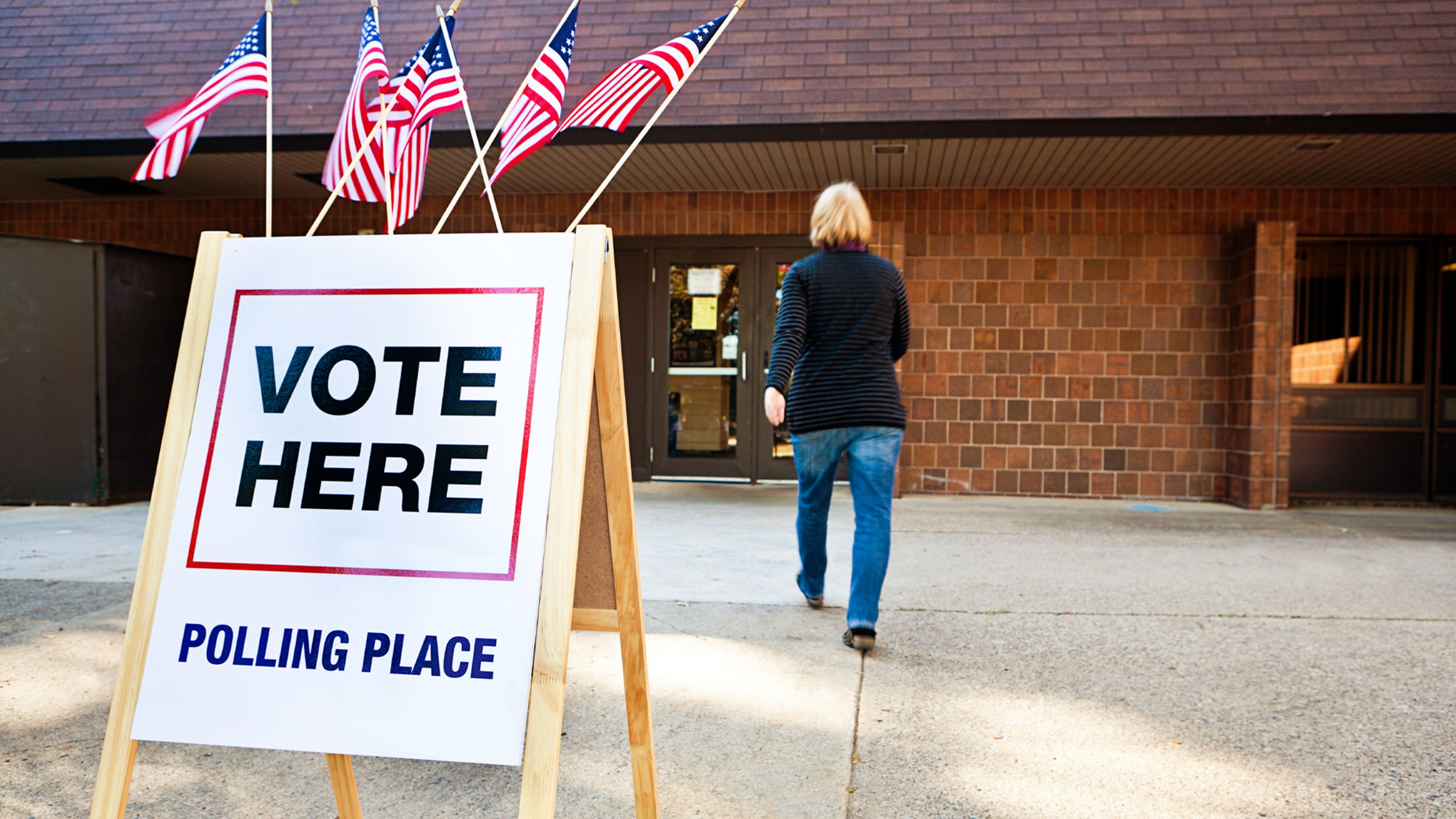 An outdoor polling place sign beside a brick building. White sign reads “VOTE HERE” and “POLLING PLACE,” with red-and-blue borders and several American flags above; a person walks toward entrance.