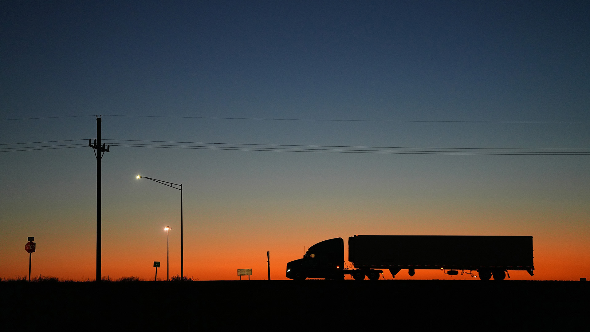 Silhouette of a semi‑truck with trailer traveling along a roadway at sunset, set against an orange and blue sky with utility poles and streetlights visible in the foreground.