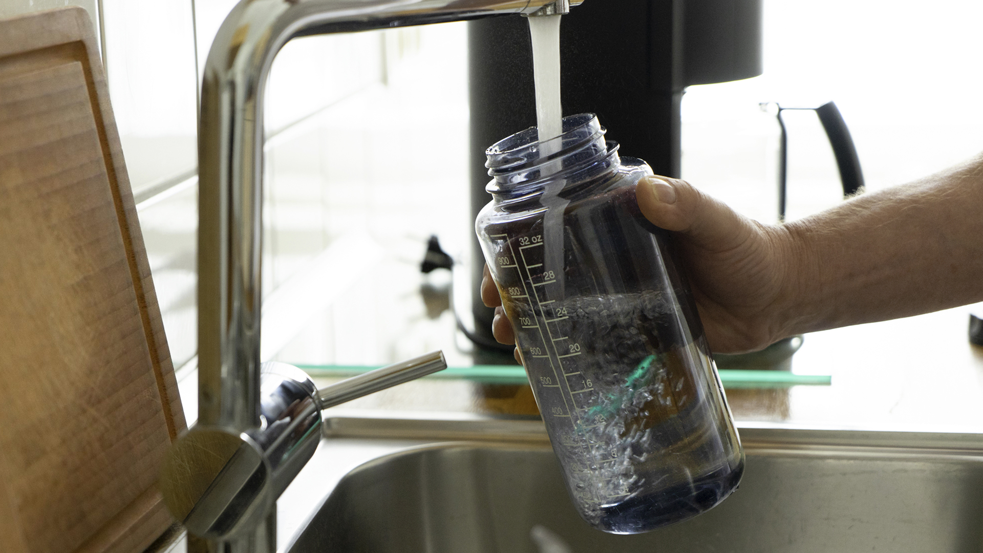A person filling a reusable transparent water bottle at a kitchen faucet above a stainless-steel sink. Bottle contains measurement markings and visible water movement, highlighting everyday tap-water use and refillable hydration.
