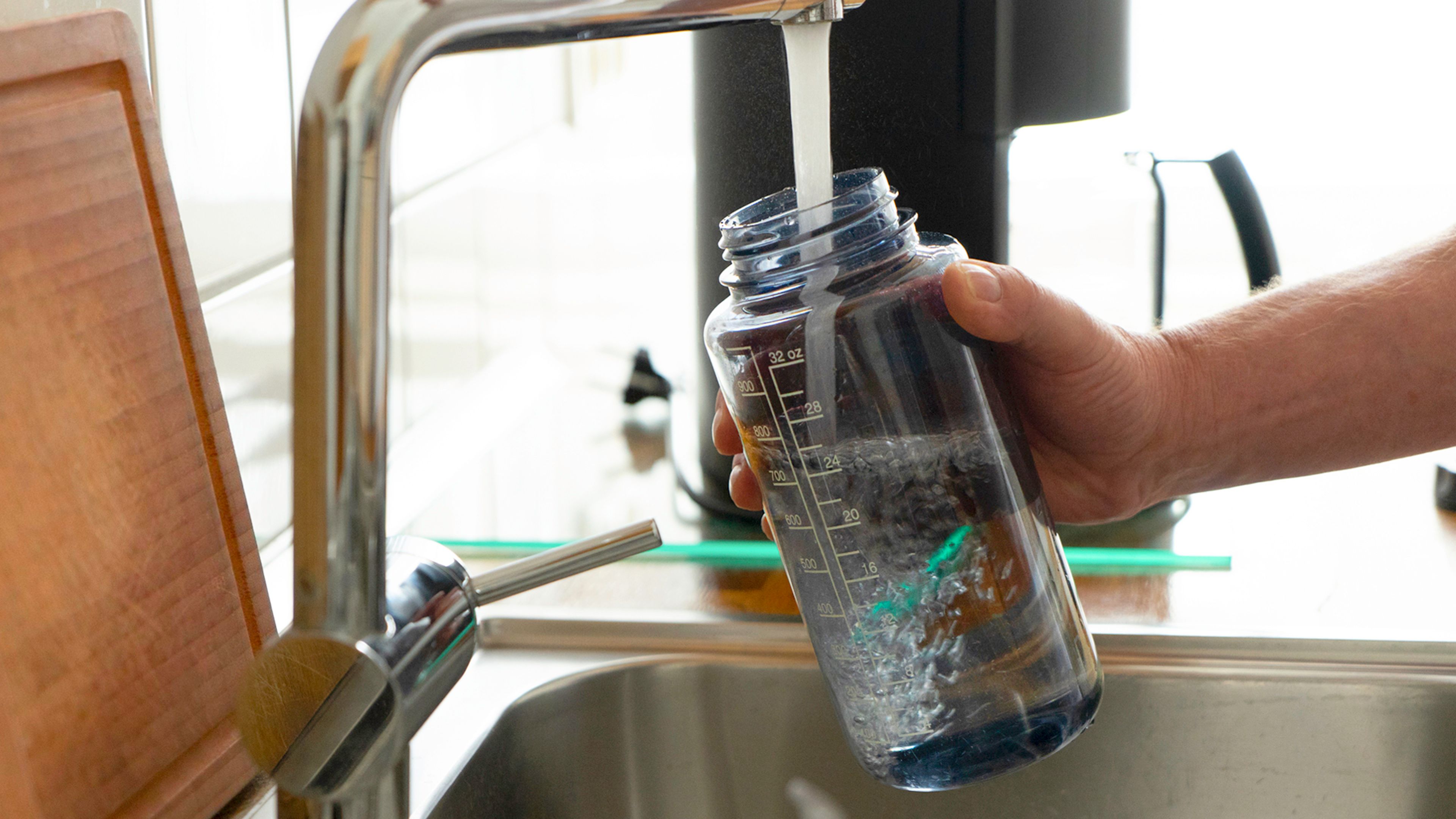 A person filling a reusable transparent water bottle at a kitchen faucet above a stainless-steel sink. Bottle contains measurement markings and visible water movement, highlighting everyday tap-water use and refillable hydration.