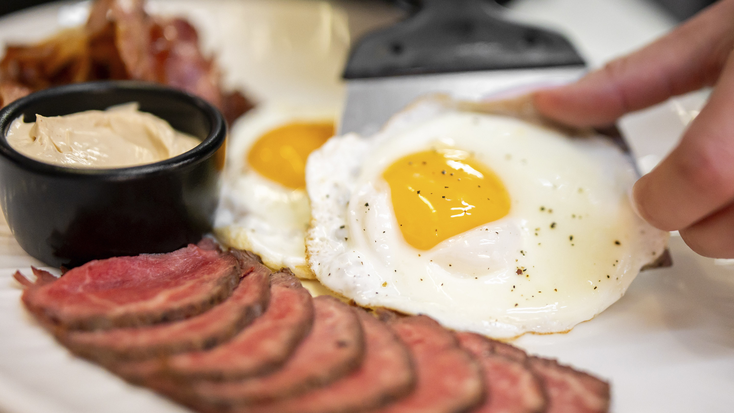 A high-angle close-up shot of a breakfast plate featuring sliced rare roast beef, sunny-side-up fried eggs, bacon, and a small bowl of sauce, with a hand adjusting an egg using a spatula
