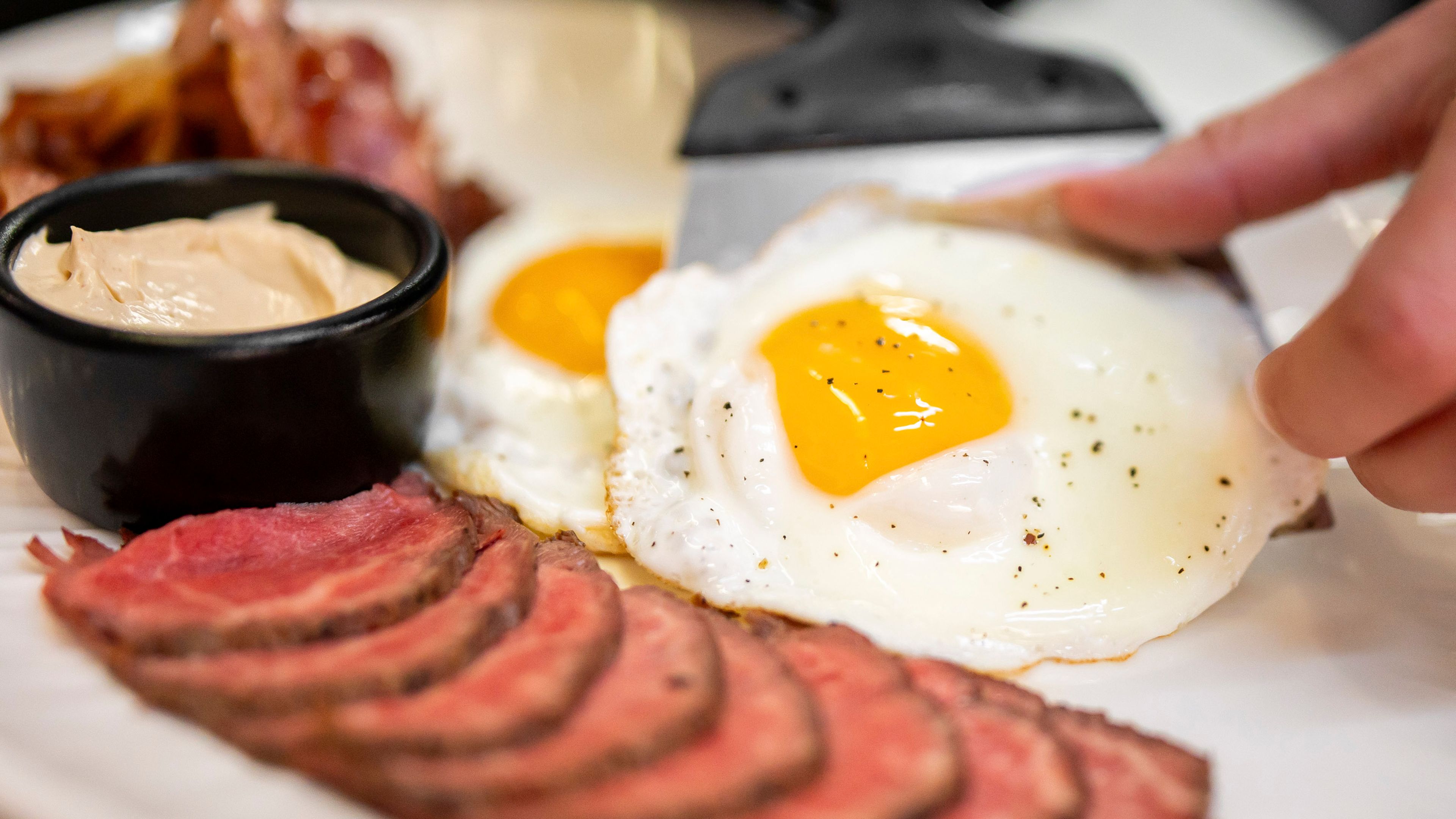 A high-angle close-up shot of a breakfast plate featuring sliced rare roast beef, sunny-side-up fried eggs, bacon, and a small bowl of sauce, with a hand adjusting an egg using a spatula