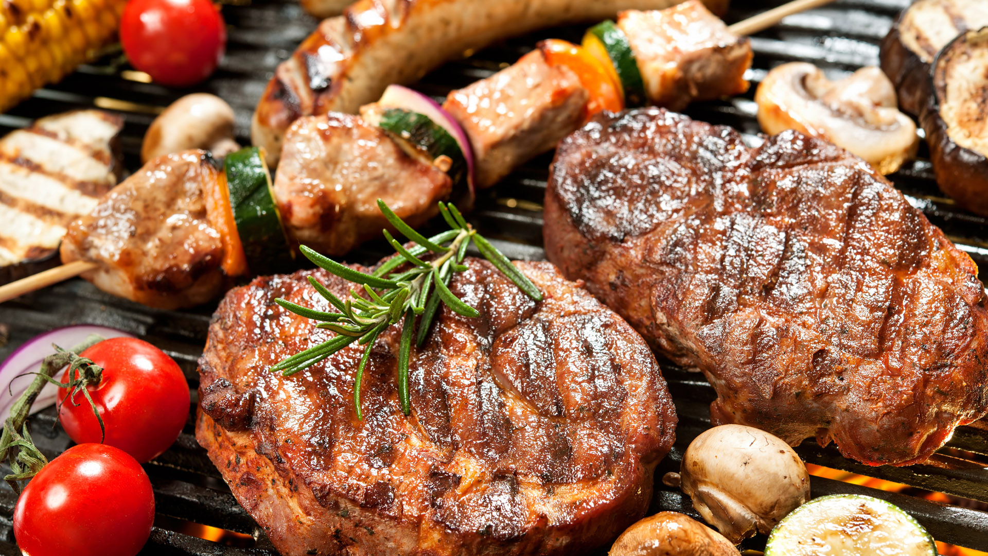 A barbecue grill with various grilled foods including two thick steaks, meat and vegetable skewers, sausages, mushrooms, cherry tomatoes, and zucchini slices. Grill marks and a sprig of rosemary on one steak highlight the cooking process and presentation of a hearty outdoor meal.