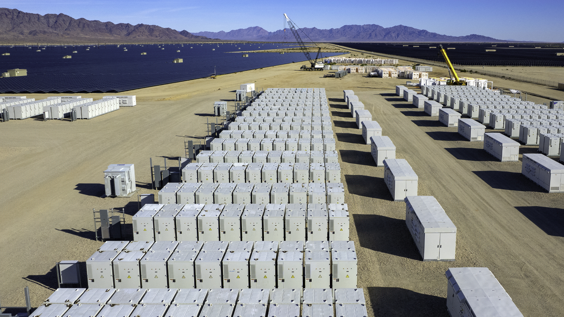 A large-scale battery energy storage facility under construction in a desert landscape beside a blue reservoir and mountain range. Long, orderly rows of white storage containers, construction cranes, access lanes, and utility equipment indicate an industrial renewable-energy project.