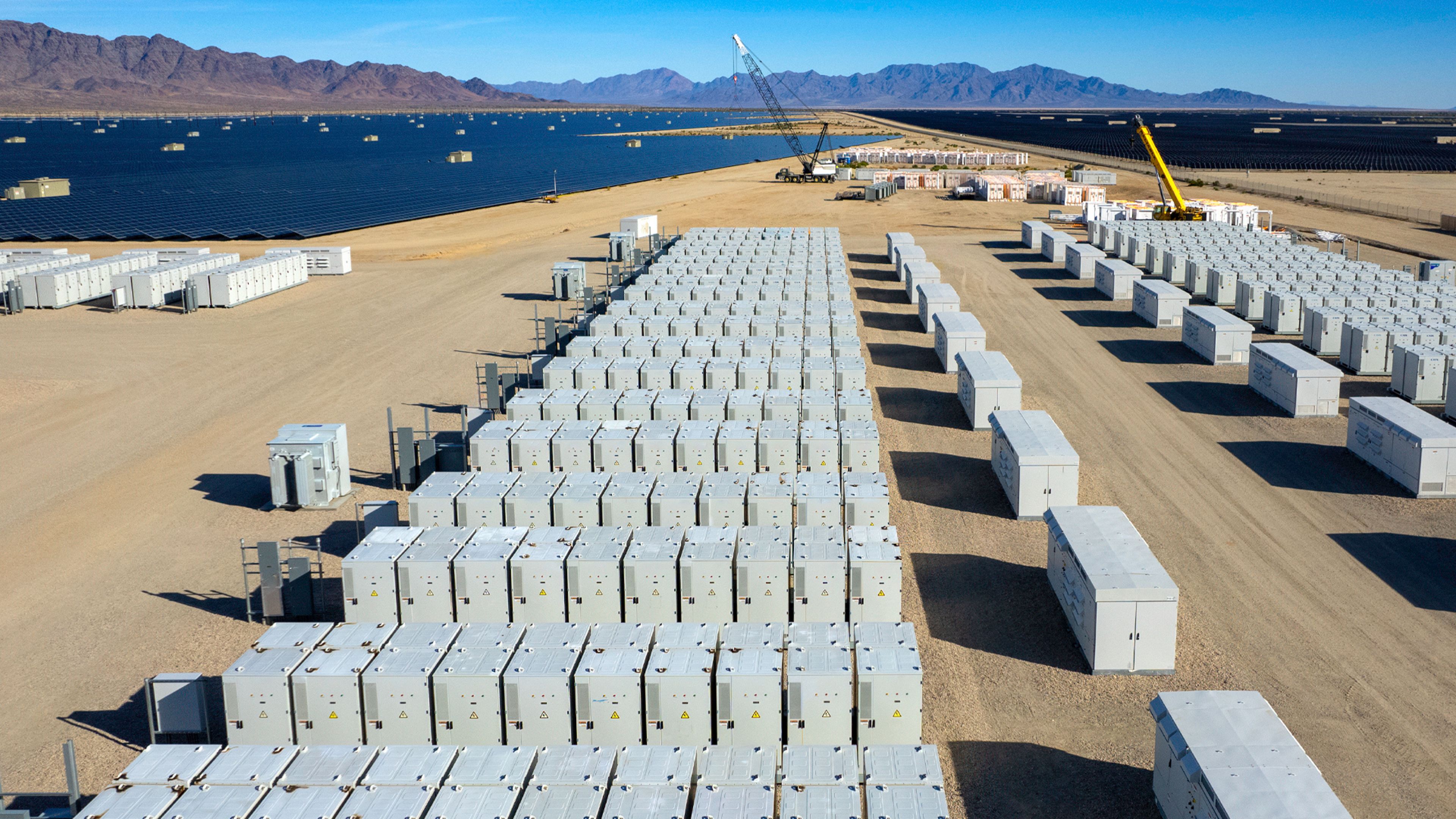 A large-scale battery energy storage facility under construction in a desert landscape beside a blue reservoir and mountain range. Long, orderly rows of white storage containers, construction cranes, access lanes, and utility equipment indicate an industrial renewable-energy project.