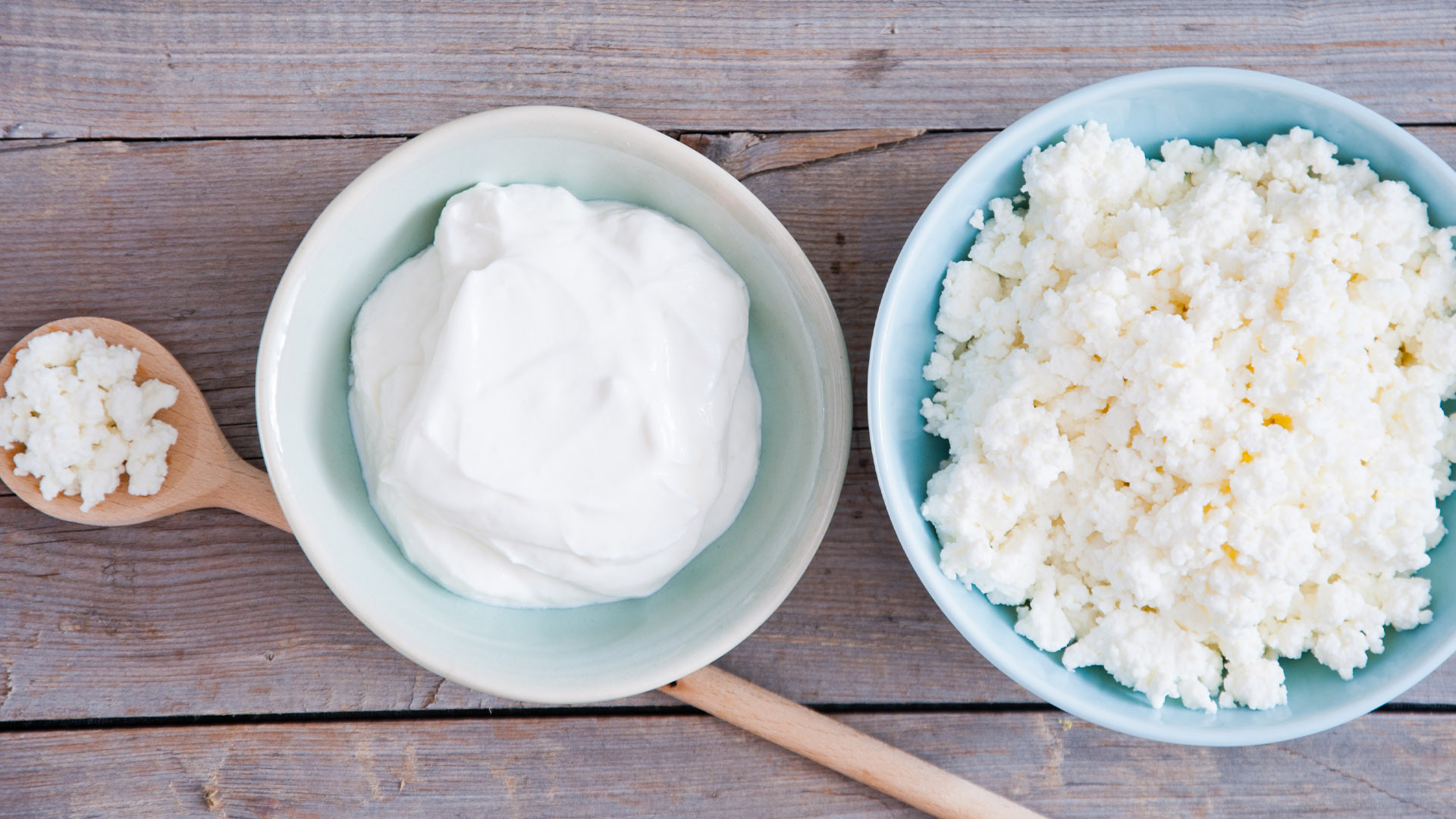 Three types of dairy products on a wooden surface: a wooden spoon with cottage cheese, a bowl with creamy yogurt, and a bowl with crumbly cottage cheese. The arrangement highlights texture differences between smooth yogurt and grainy cottage cheese.