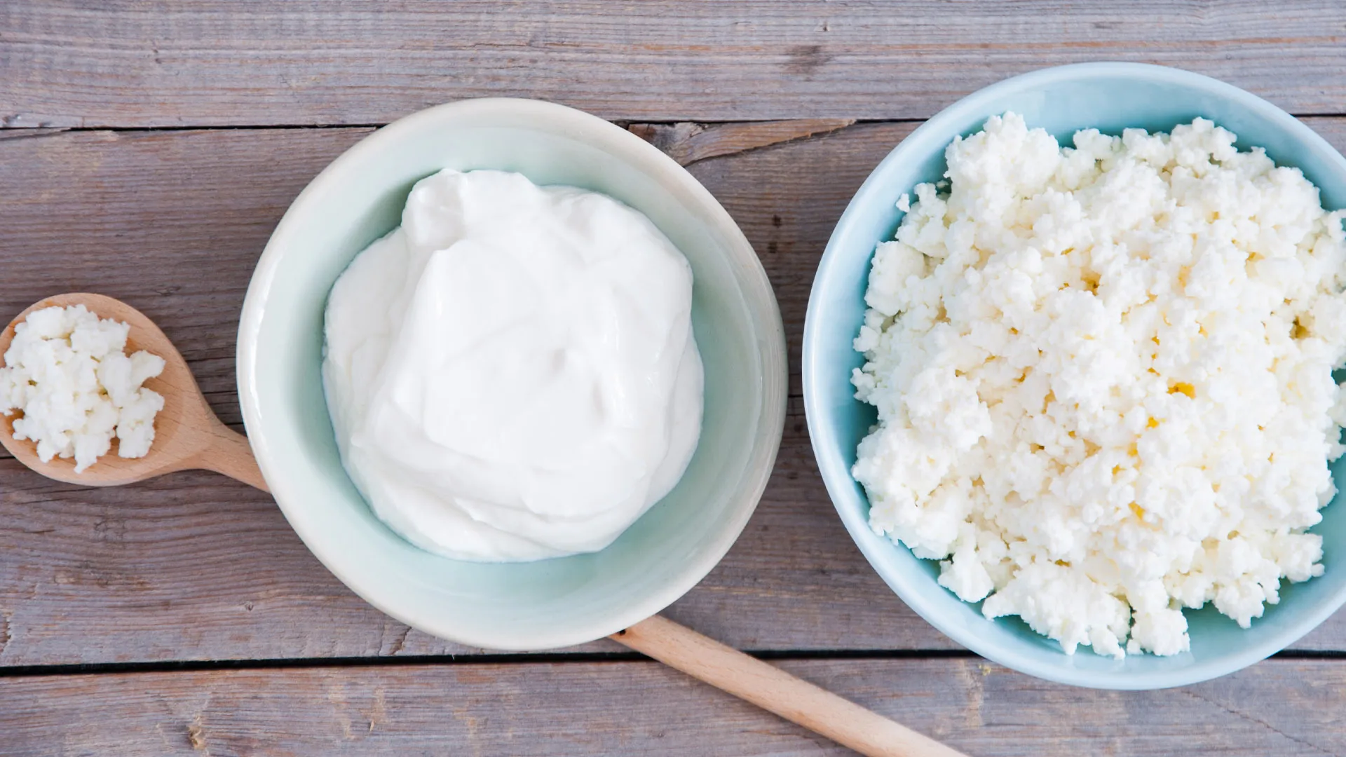 Three types of dairy products on a wooden surface: a wooden spoon with cottage cheese, a bowl with creamy yogurt, and a bowl with crumbly cottage cheese. The arrangement highlights texture differences between smooth yogurt and grainy cottage cheese.