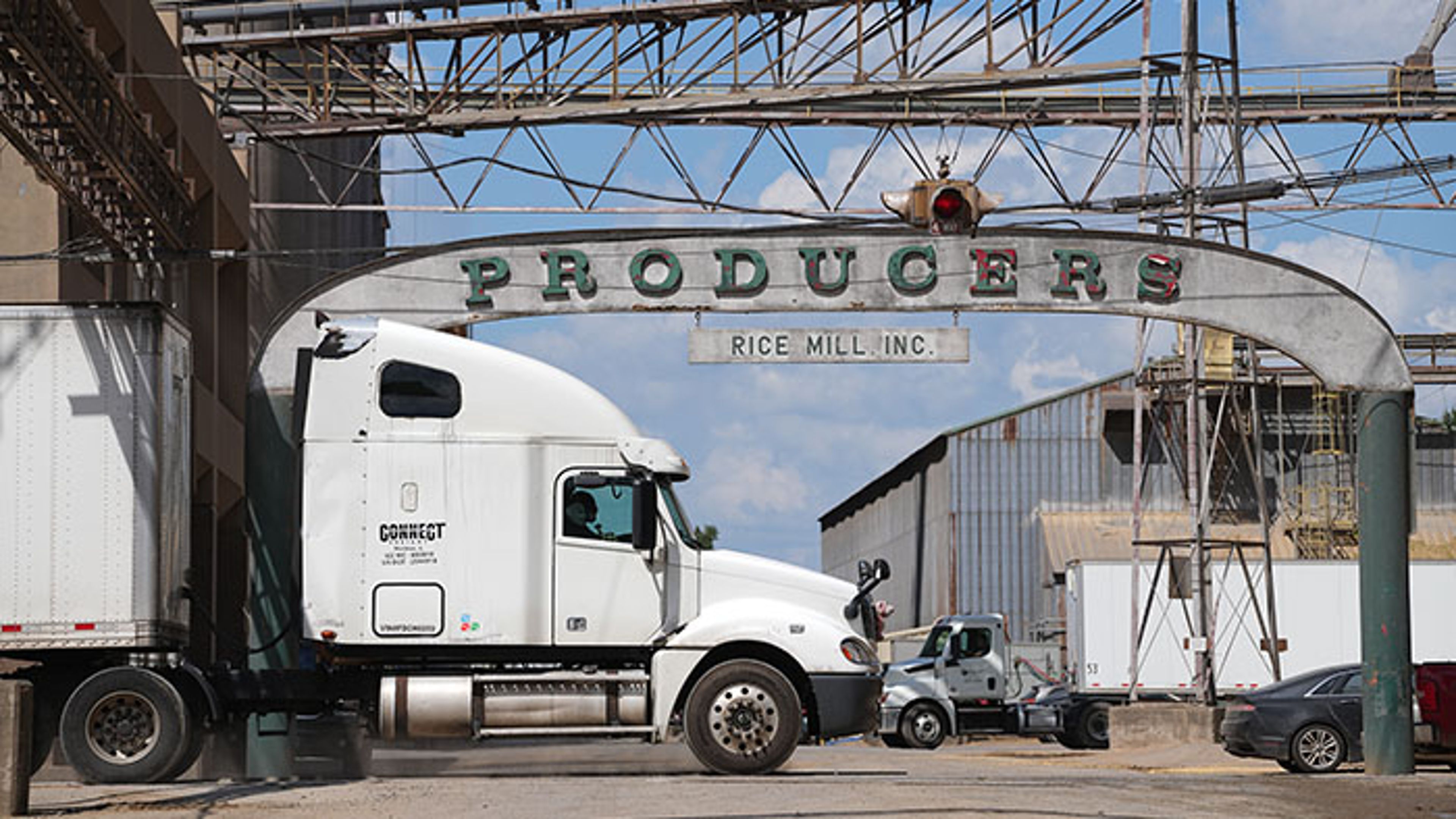 Truck hauling grain trailer passing beneath pipe conveyor system at a rice mill facility