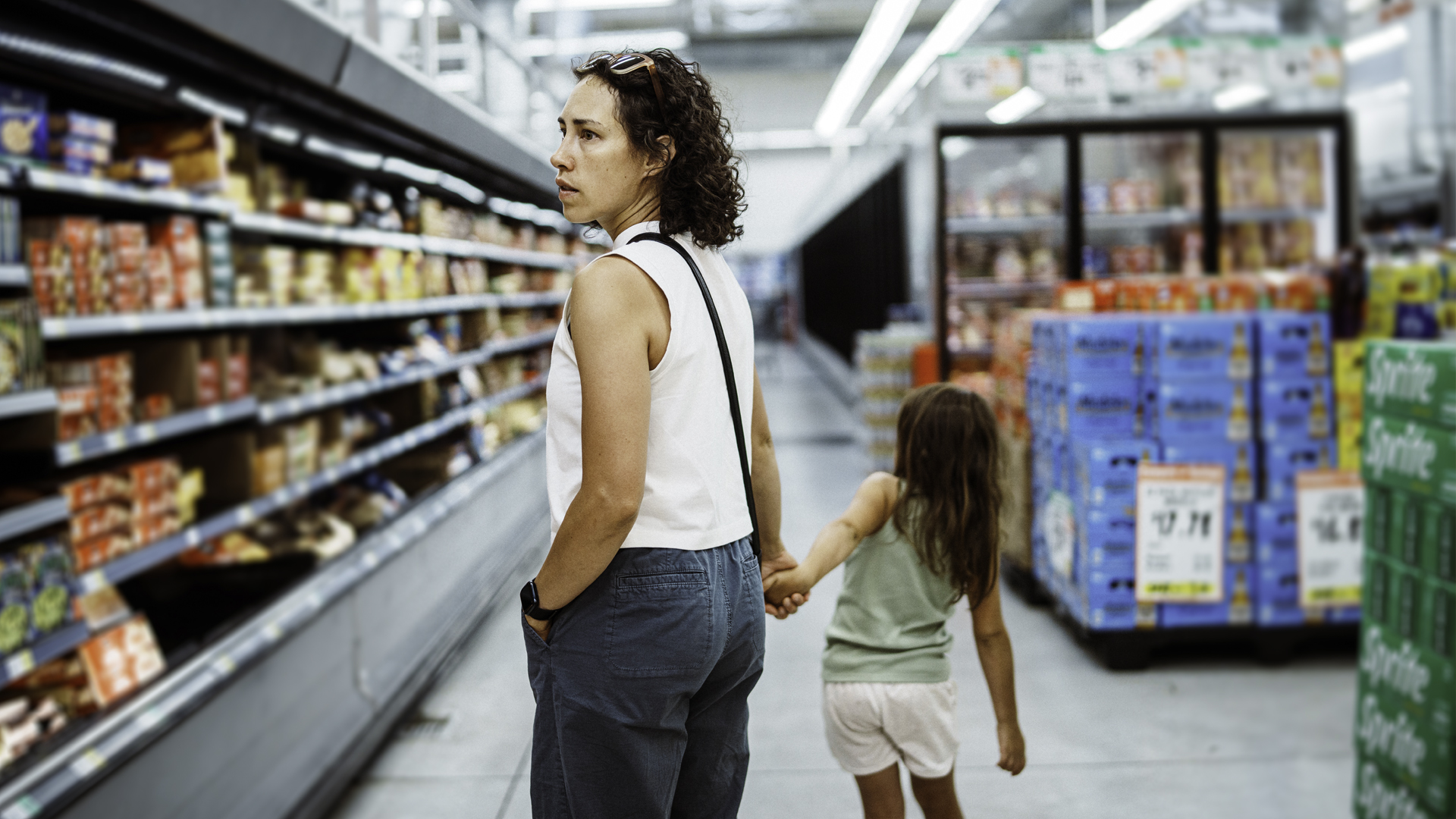 Person shopping in a grocery store aisle while holding a child’s hand, with shelves of food products and beverage displays in the background.