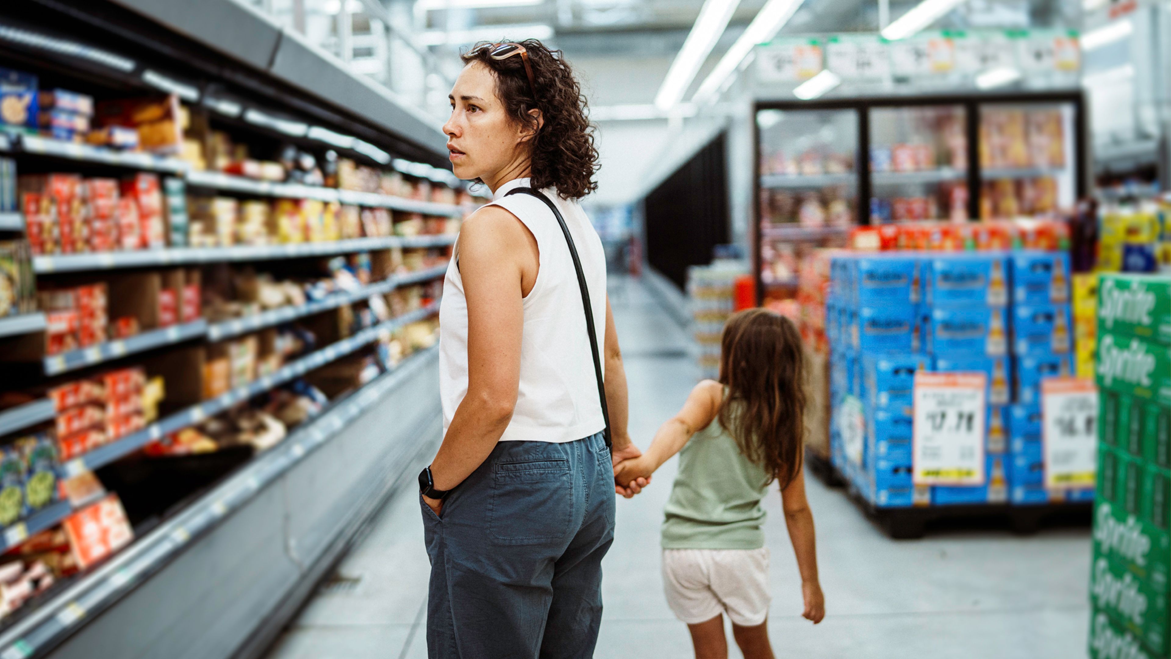Person shopping in a grocery store aisle while holding a child’s hand, with shelves of food products and beverage displays in the background.