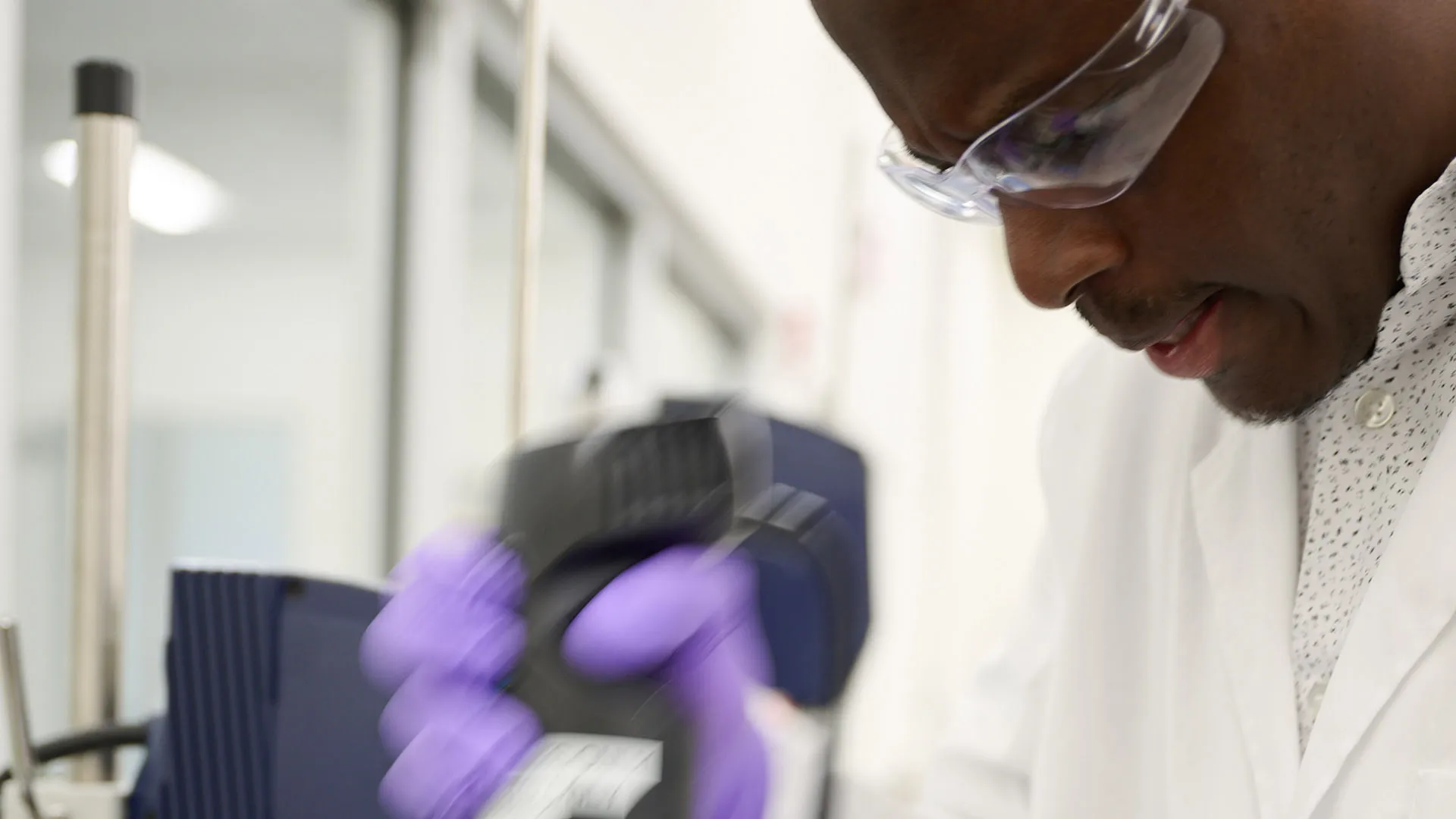 A laboratory worker in a white coat using handheld equipment near scientific instruments.