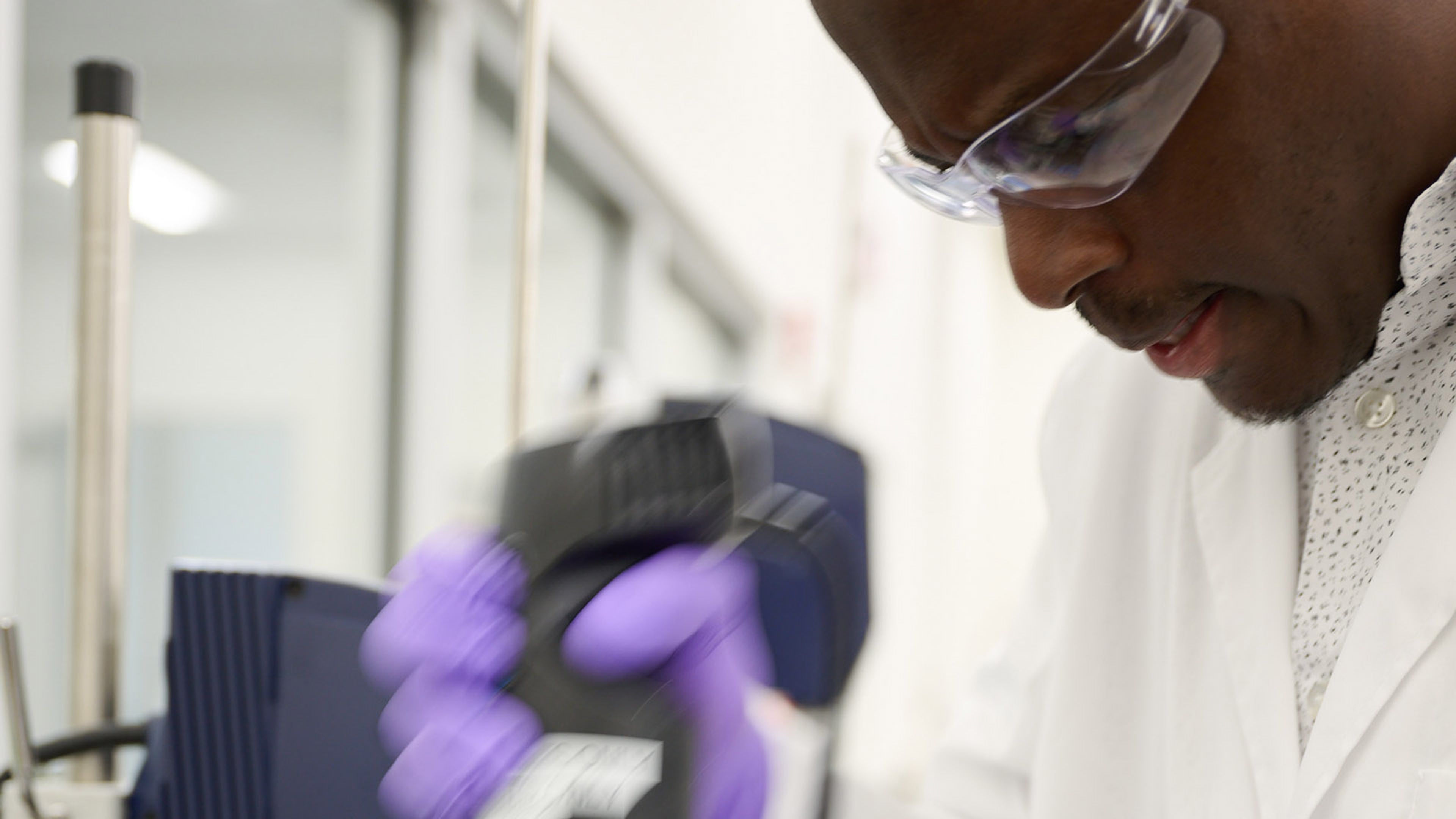 A laboratory worker in a white coat using handheld equipment near scientific instruments.