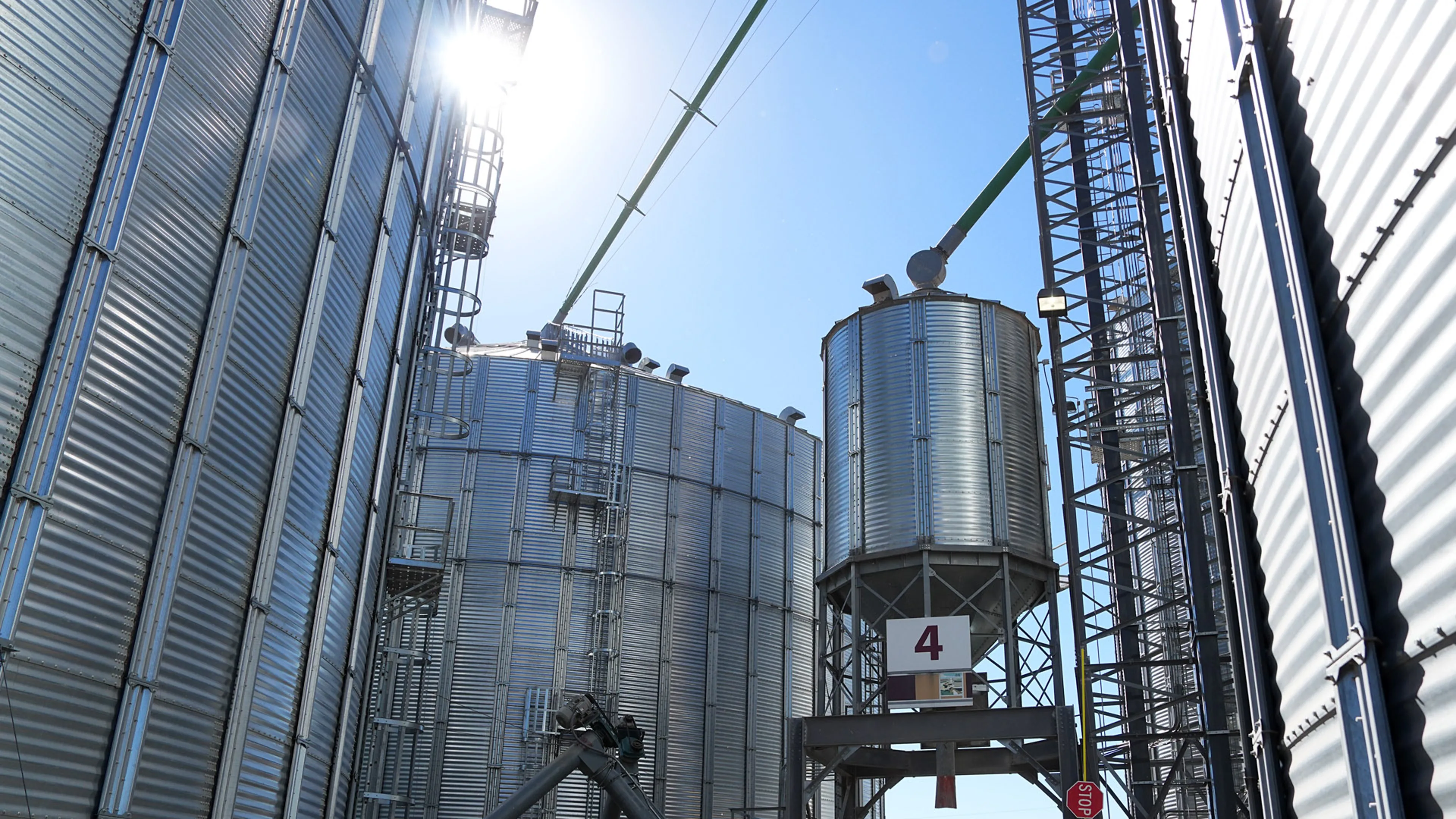 Tall metal grain storage bins and towers at an agricultural complex with sunlight shining through.