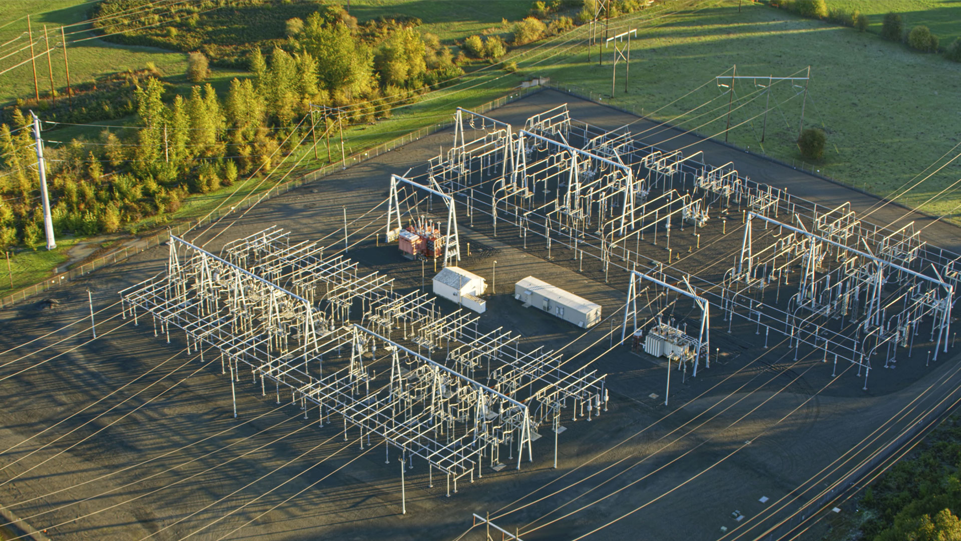 Aerial view of an electrical substation with numerous metal frameworks, transformers, and power lines arranged in a grid pattern, surrounded by green fields and trees.