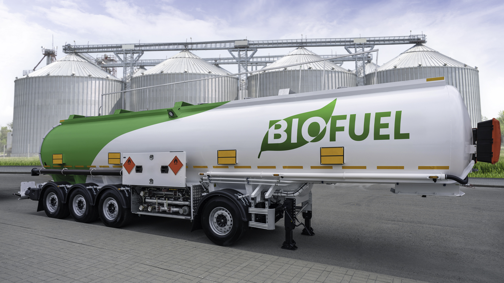 A biofuel tanker truck parked beside large industrial grain silos. White-and-green cylindrical trailer displays prominent “BIOFUEL” branding, with multiple axles, hazard placards, amber marker lights, hoses, and rear equipment visible.