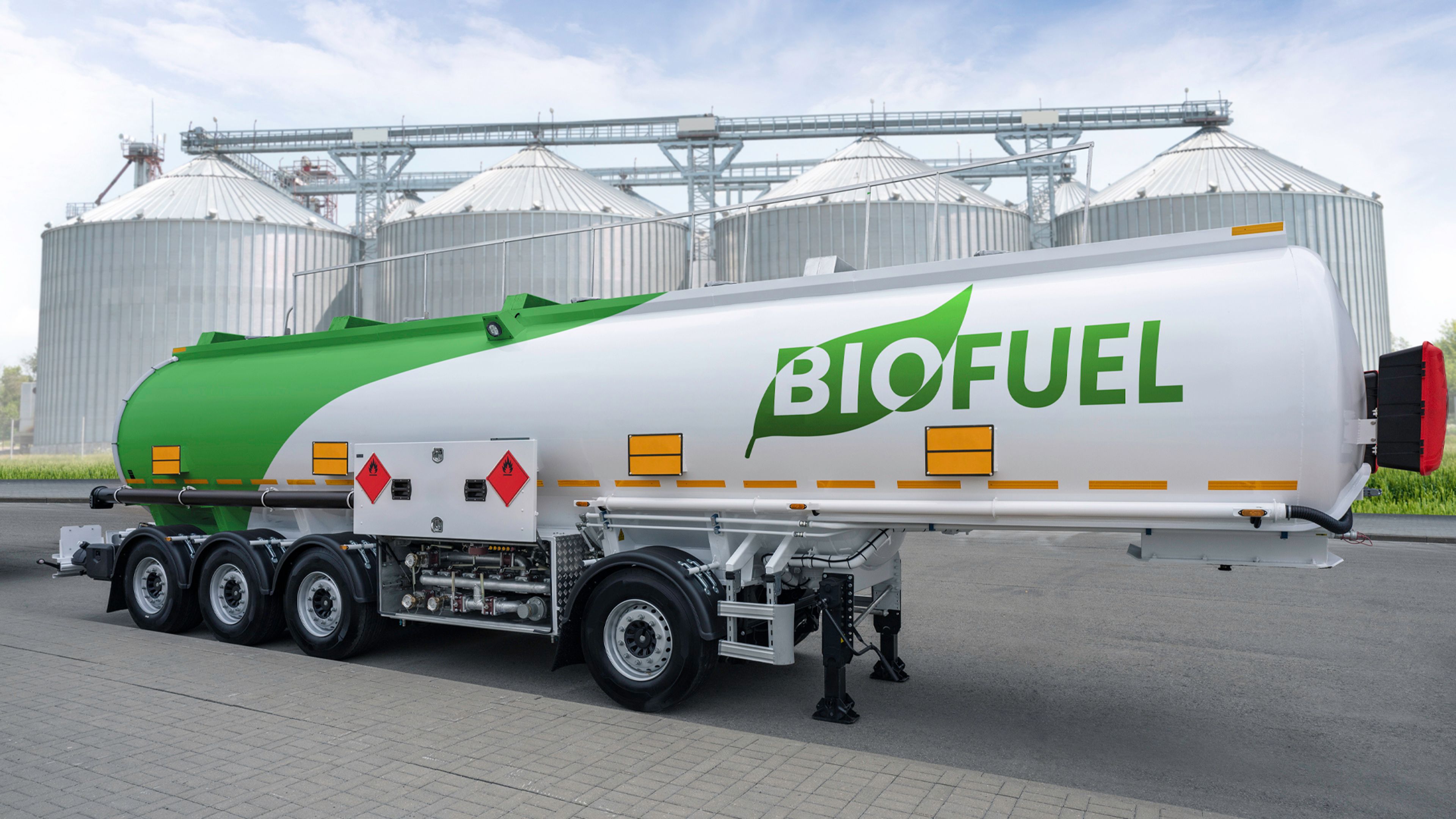 A biofuel tanker truck parked beside large industrial grain silos. White-and-green cylindrical trailer displays prominent “BIOFUEL” branding, with multiple axles, hazard placards, amber marker lights, hoses, and rear equipment visible.