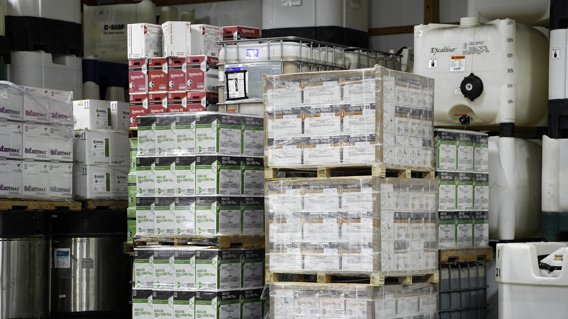 Pallets of agricultural fertilizer and crop nutrient products stored in a warehouse, including boxed and liquid formulations stacked on shelving and in bulk containers.