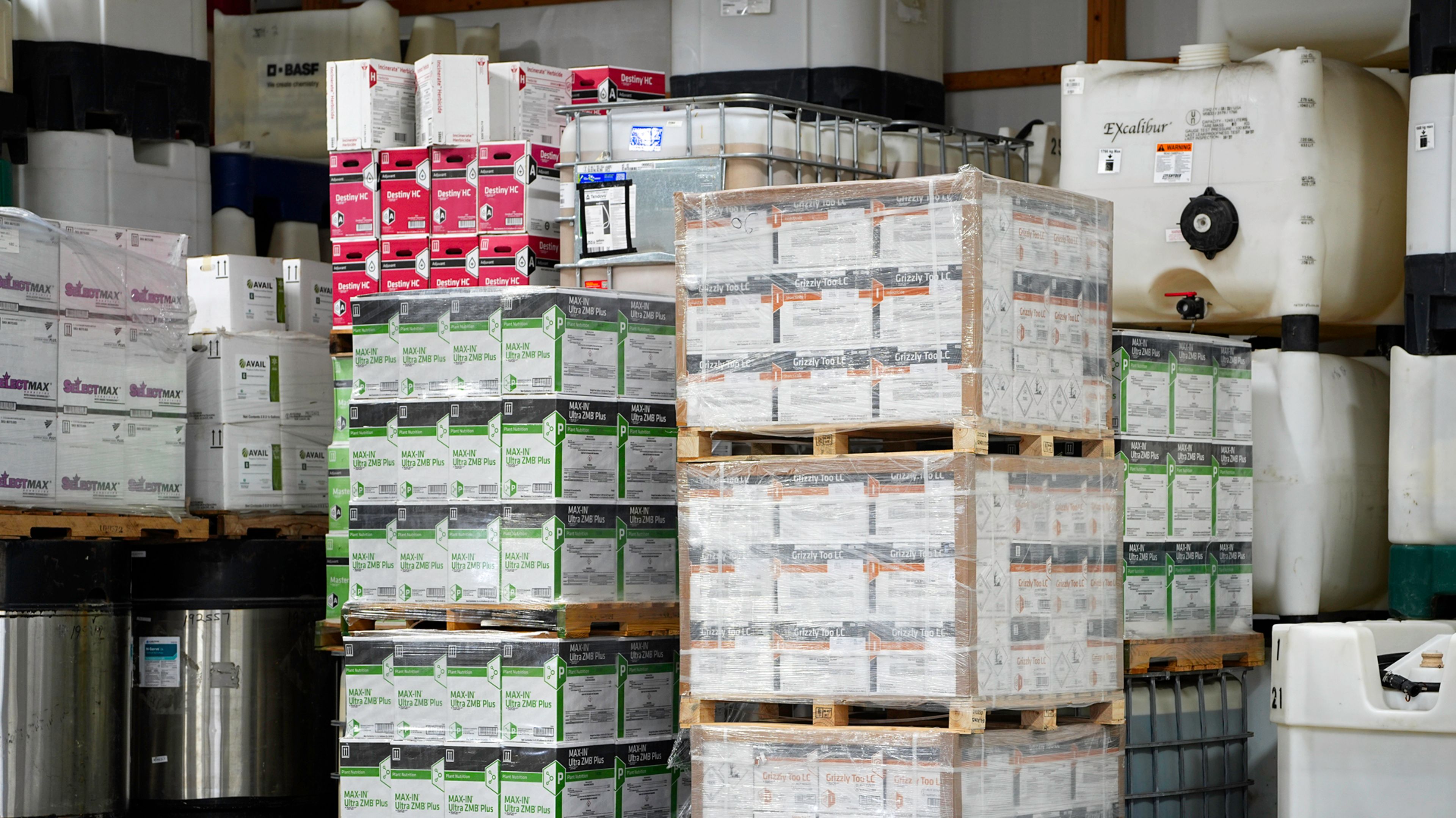 Pallets of agricultural fertilizer and crop nutrient products stored in a warehouse, including boxed and liquid formulations stacked on shelving and in bulk containers.