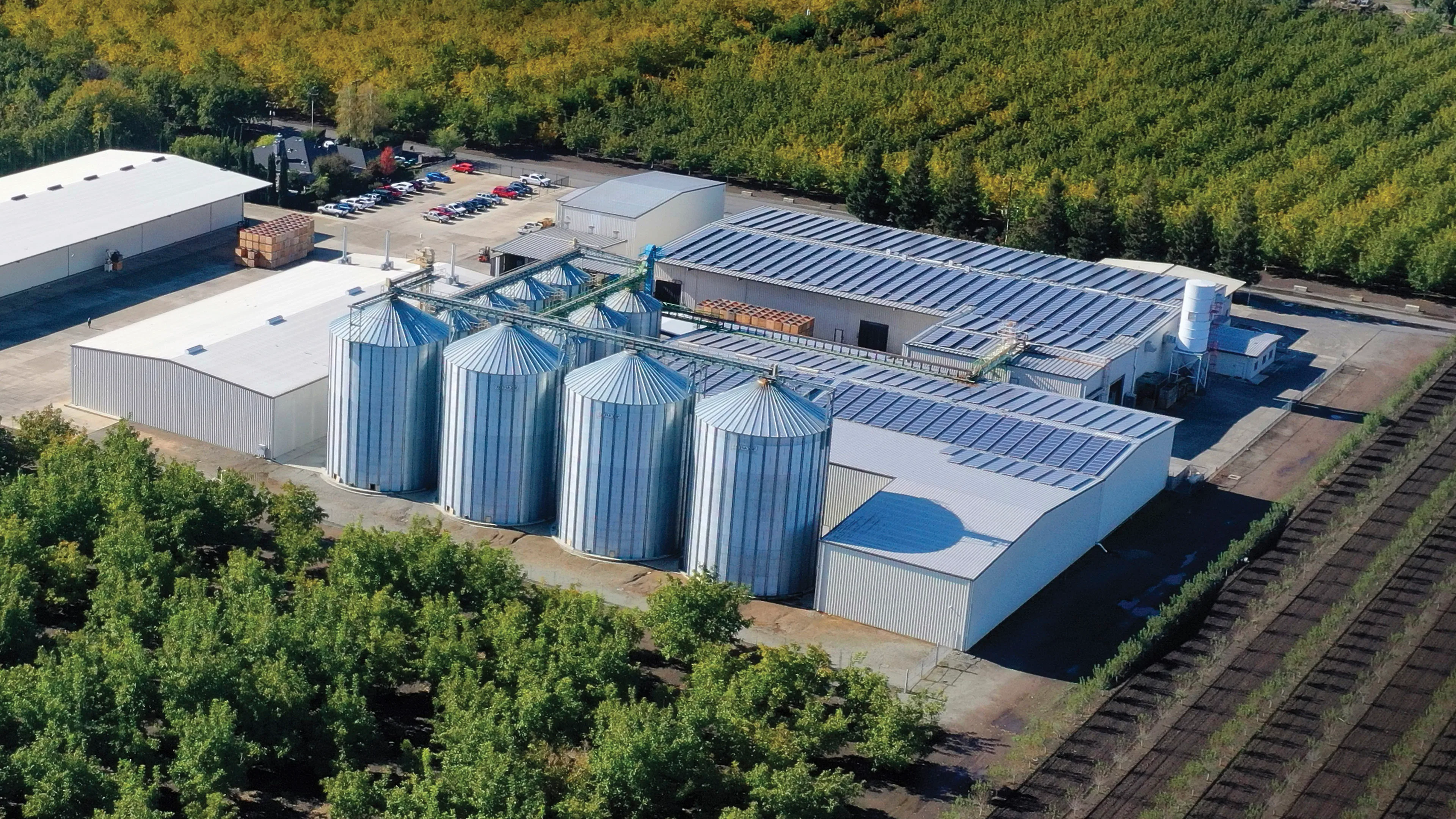 Aerial view of an agricultural processing facility with multiple metal grain silos connected to large warehouse buildings with rooftop solar panels, surrounded by orchards and a small parking lot.