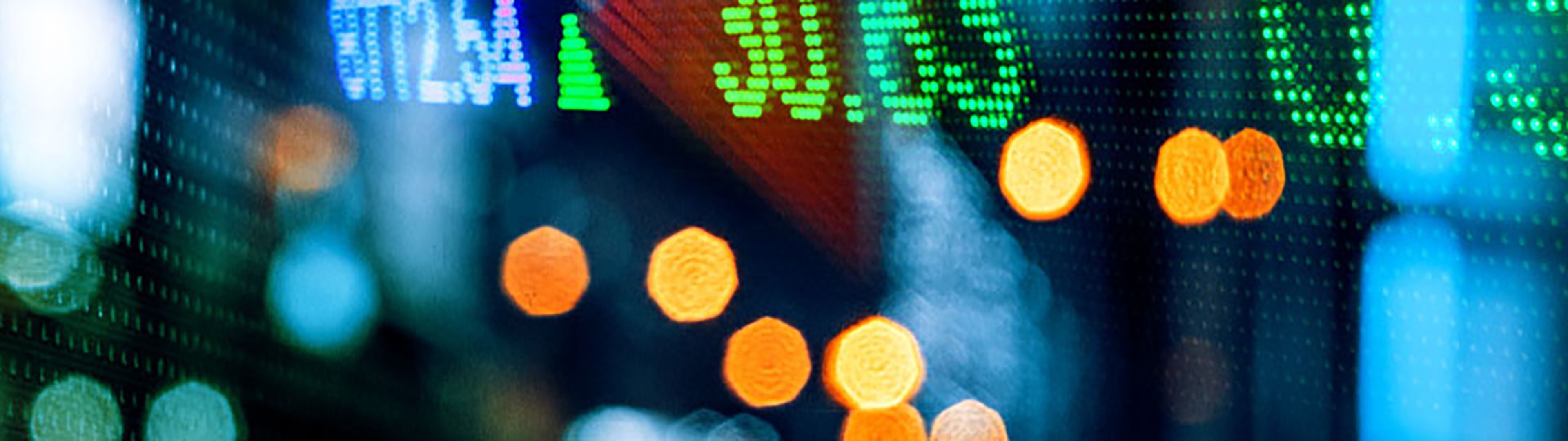 Blurred close-up of a digital stock market display with green numeric indicators and arrows, including upward and downward trends, overlaid with out-of-focus orange and gold lights in the foreground, creating an abstract financial market scene.