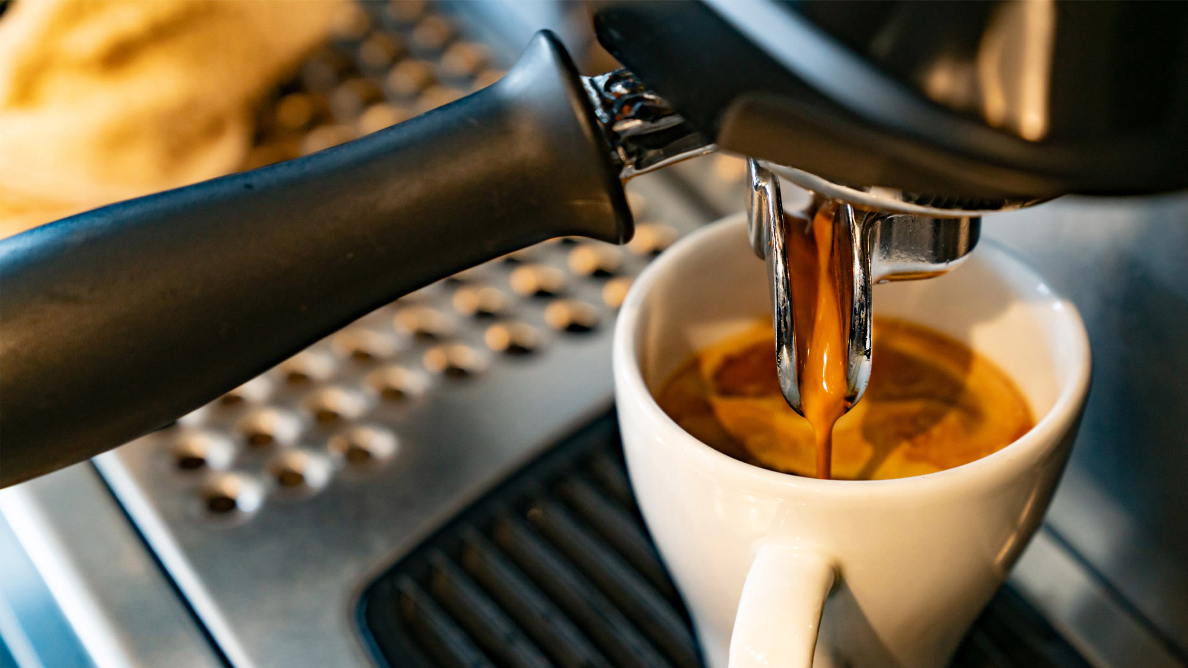 An espresso being brewed from a portafilter into a white ceramic cup on a metal drip tray. Rich, dark coffee flows smoothly, highlighting the brewing process and crema forming on top.