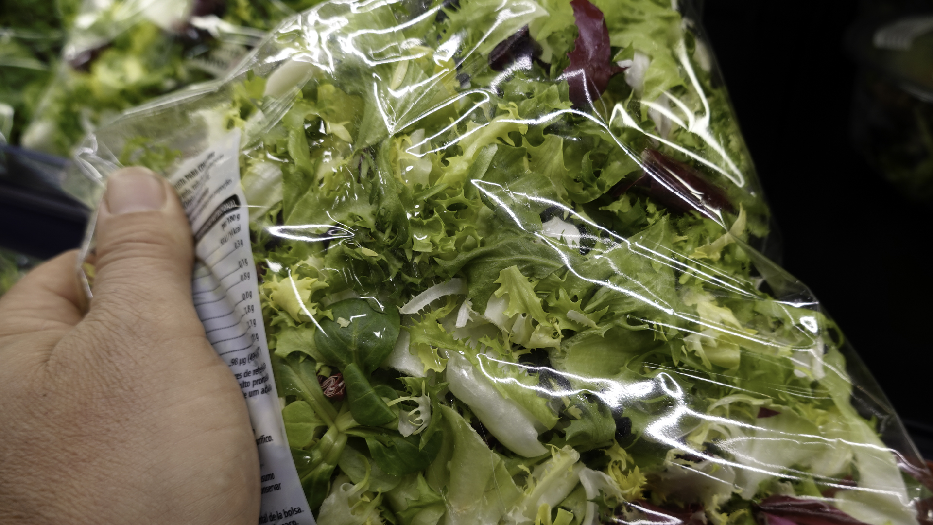 A packaged mixed green salad held by a hand in a grocery setting. Clear plastic packaging reveals assorted lettuce, leafy greens, pale cabbage, and red cabbage, alongside a partially visible nutrition label.