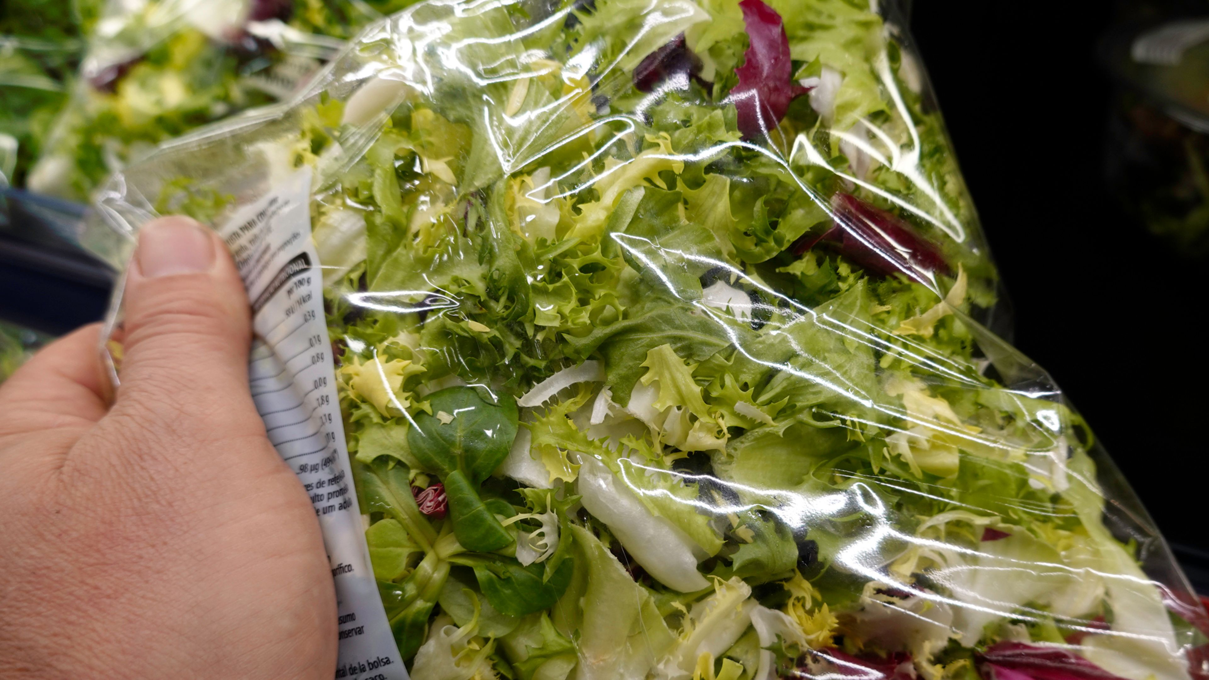 A packaged mixed green salad held by a hand in a grocery setting. Clear plastic packaging reveals assorted lettuce, leafy greens, pale cabbage, and red cabbage, alongside a partially visible nutrition label.