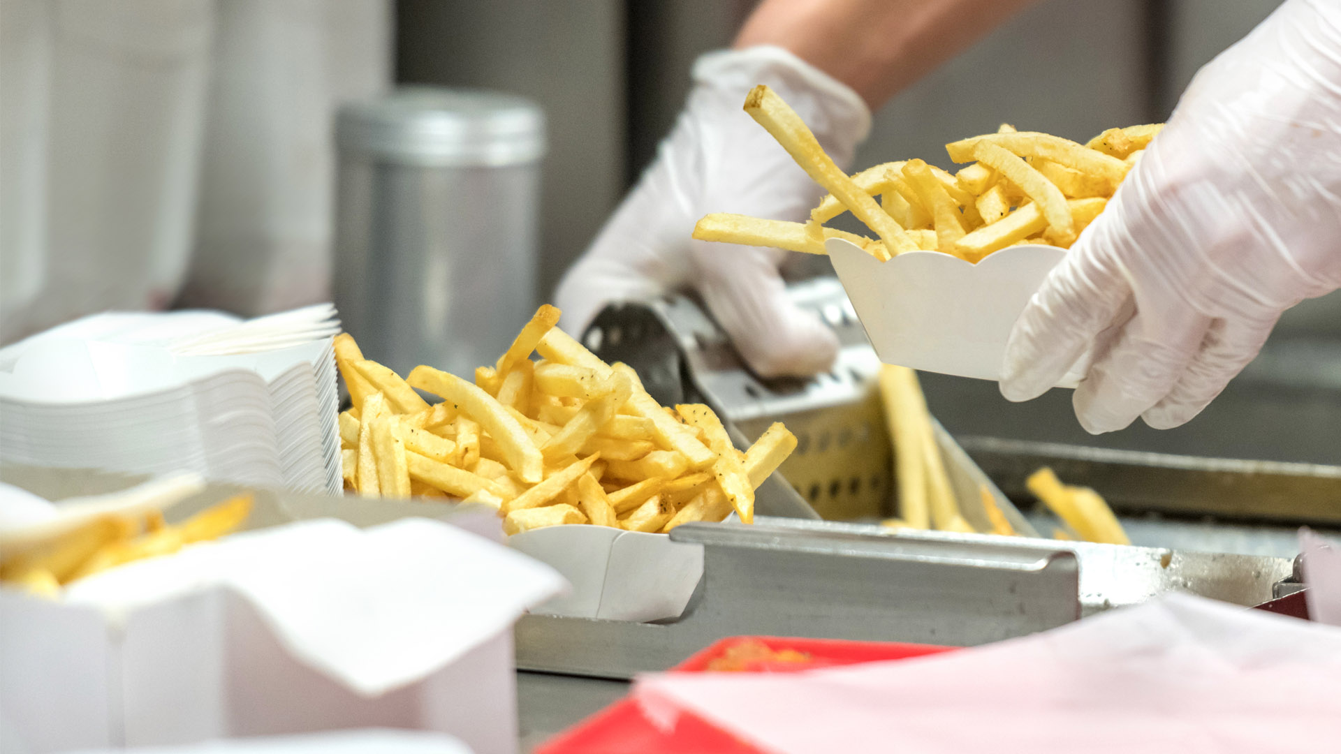 A close-up of a person wearing gloves scooping cooked French fries into a white paper container at a food service station. The scene highlights food preparation with multiple containers filled with fries and a metal tray in the background.