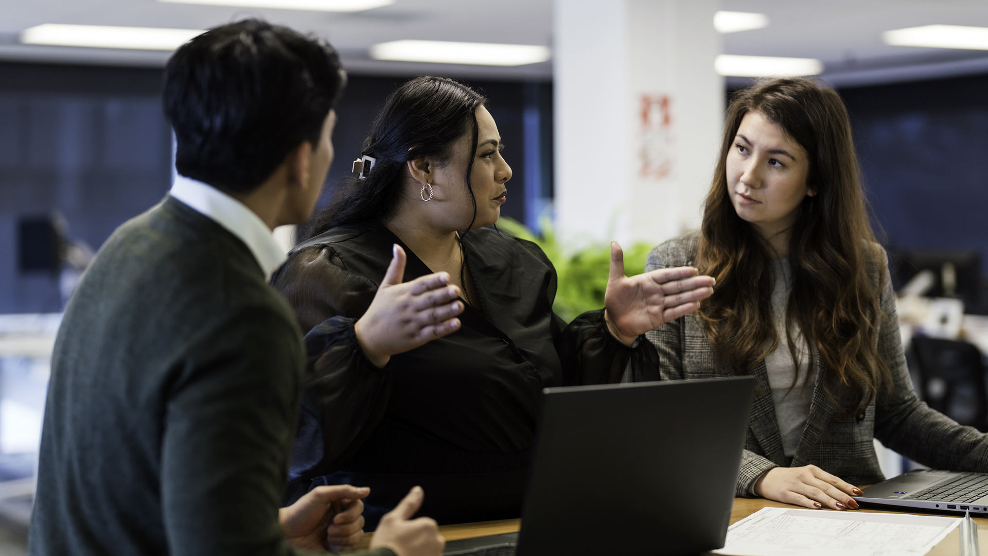 Three colleagues collaborate around a table in an office setting. One person gestures with both hands while speaking, as two others listen and look toward a laptop and documents spread across the table. The workspace features modern lighting and a blurred office background.