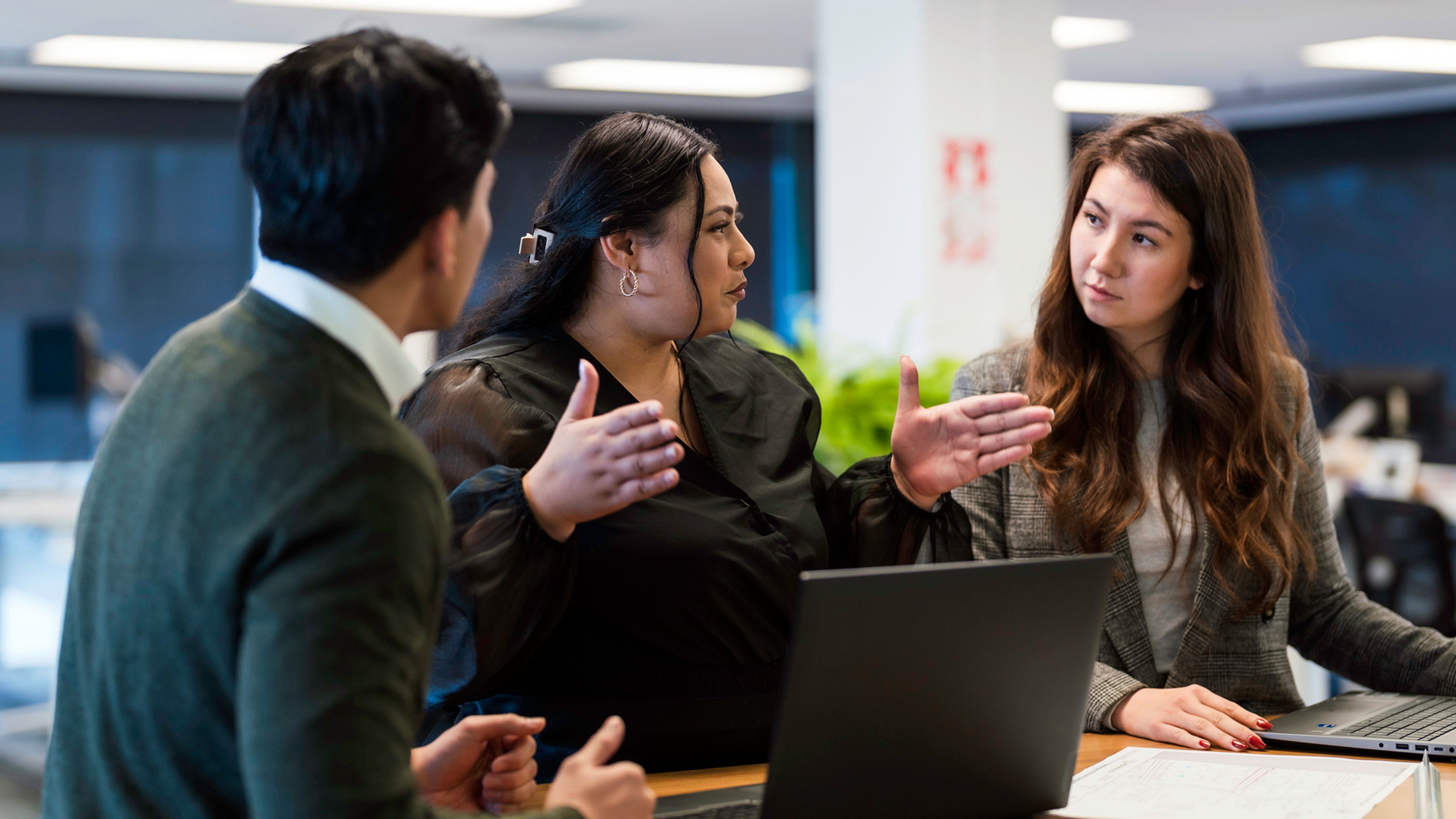 Three colleagues collaborate around a table in an office setting. One person gestures with both hands while speaking, as two others listen and look toward a laptop and documents spread across the table. The workspace features modern lighting and a blurred office background.