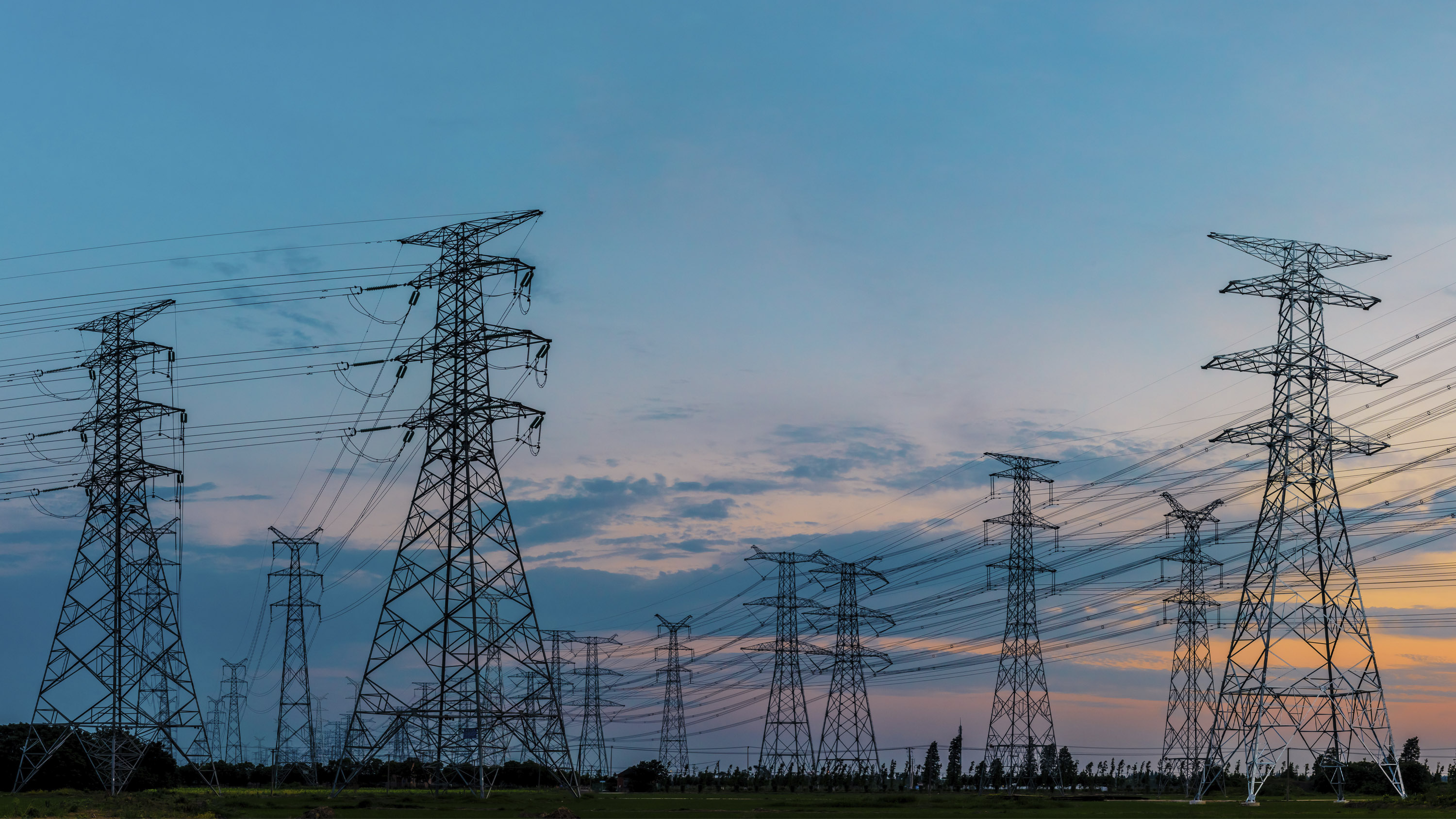 High-voltage electrical transmission towers and power lines stretching across an open landscape beneath a blue and pink sunset sky.