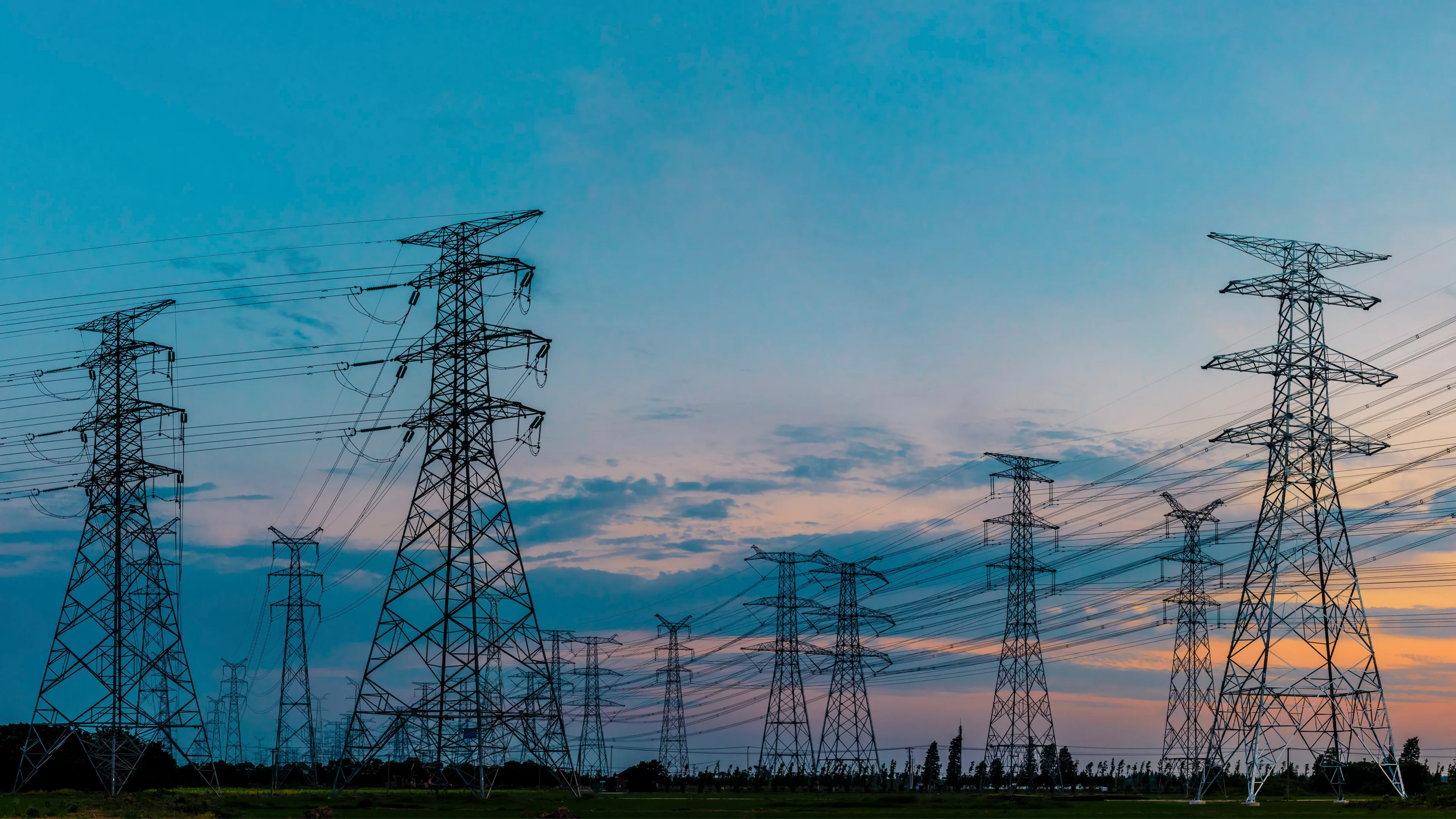 High-voltage electrical transmission towers and power lines stretching across an open landscape beneath a blue and pink sunset sky.