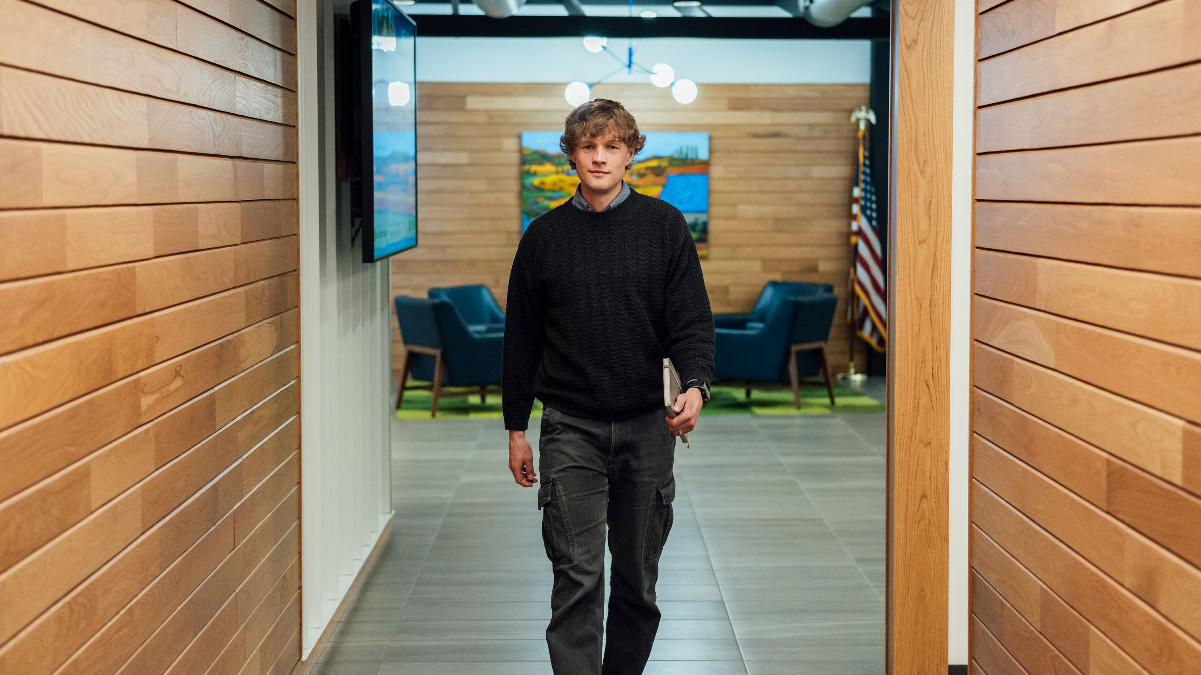 Person walking down a hallway between wood-paneled walls toward an open office space with seating and screens in the background