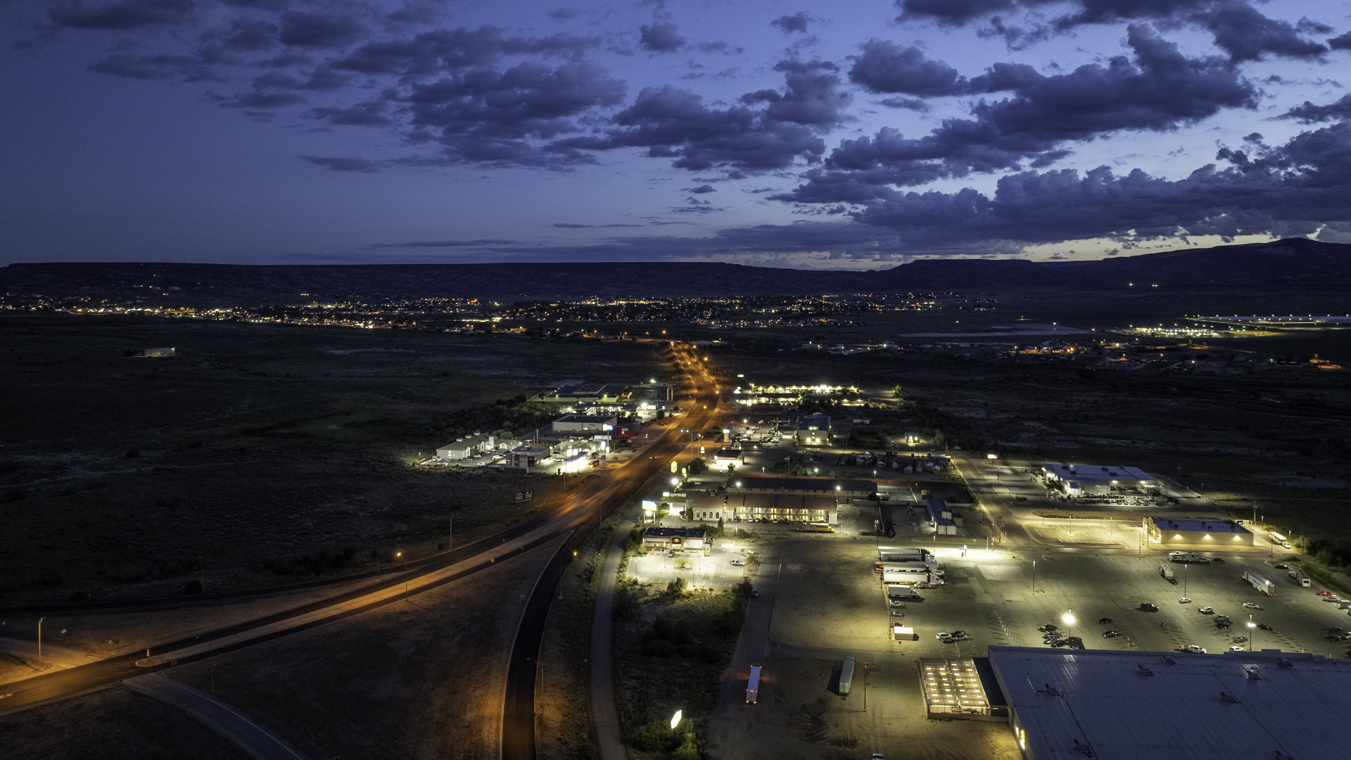 Aerial photograph shows a town and commercial district at dusk, highlighting illuminated roads, parking lots, buildings, and surrounding dark landscape. Orange streetlights lead toward distant settlements and mountains beneath a blue, cloud-filled sky.