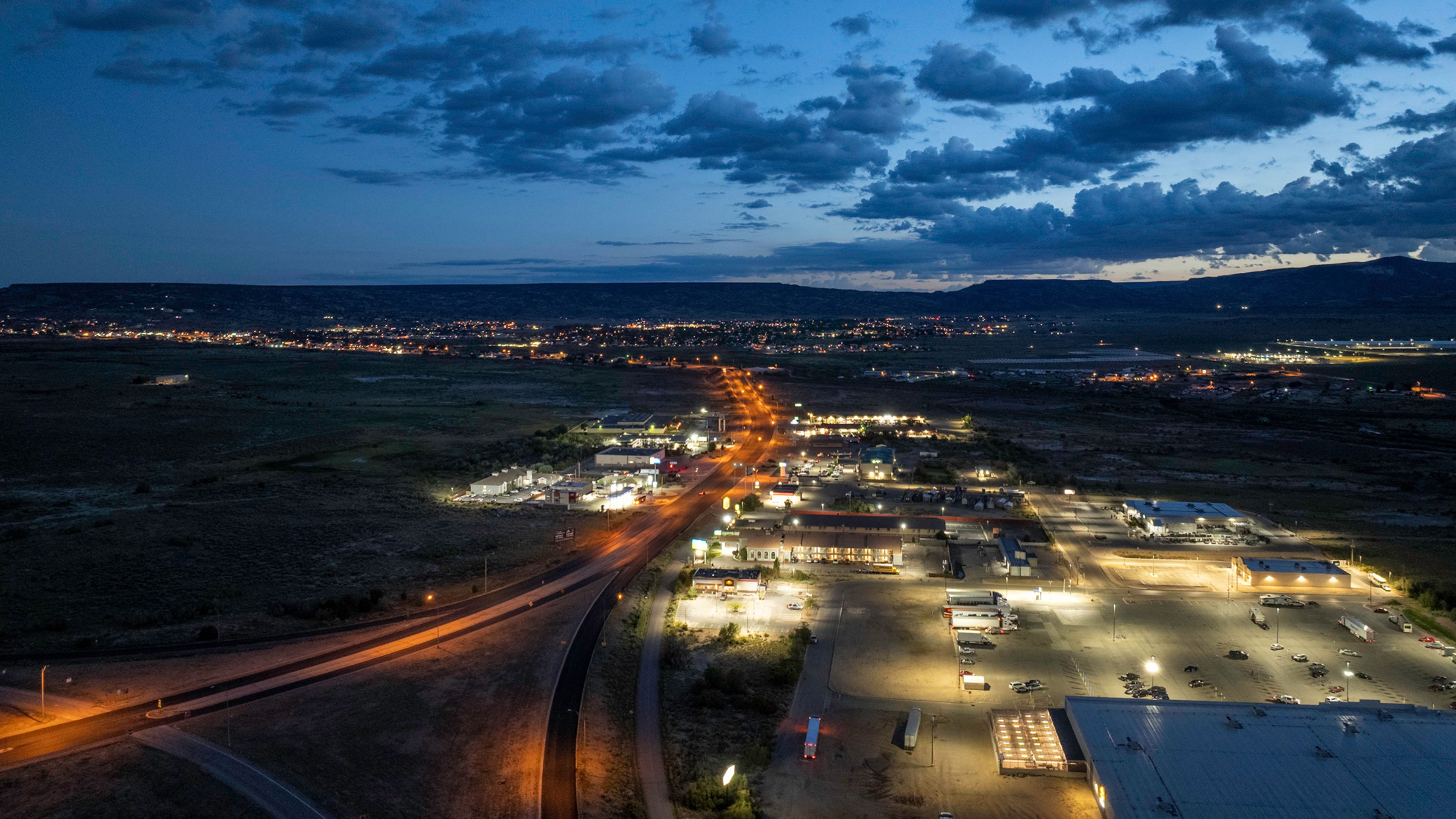 Aerial photograph shows a town and commercial district at dusk, highlighting illuminated roads, parking lots, buildings, and surrounding dark landscape. Orange streetlights lead toward distant settlements and mountains beneath a blue, cloud-filled sky.