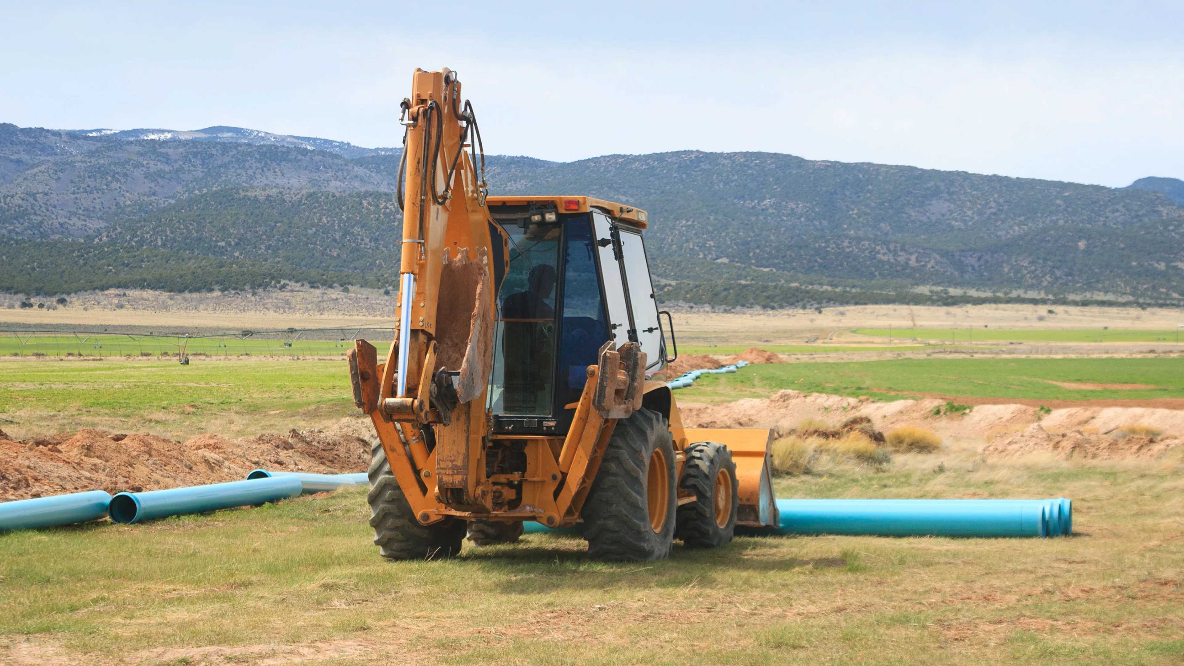 Construction equipment positioned in a field near installed piping, with open land and distant mountains in the background