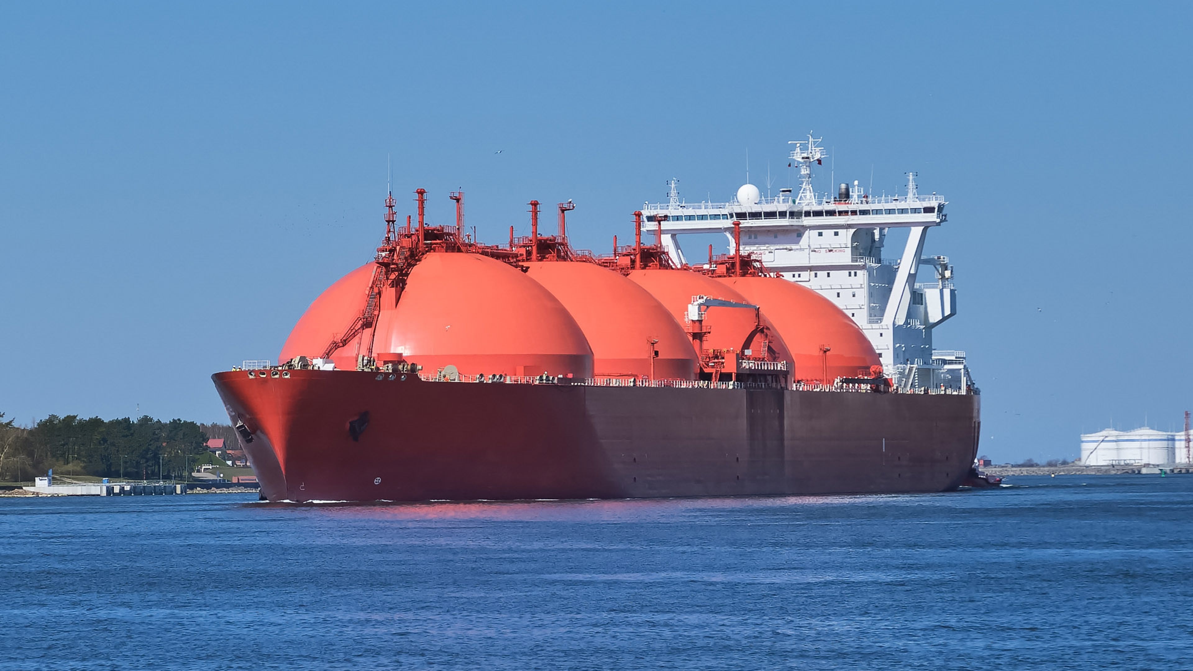 A large liquefied natural gas (LNG) carrier ship with three prominent orange spherical tanks on deck, floating on calm water near a shoreline. The ship is designed for transporting LNG, with visible infrastructure for loading and unloading gas, set against a clear blue sky.