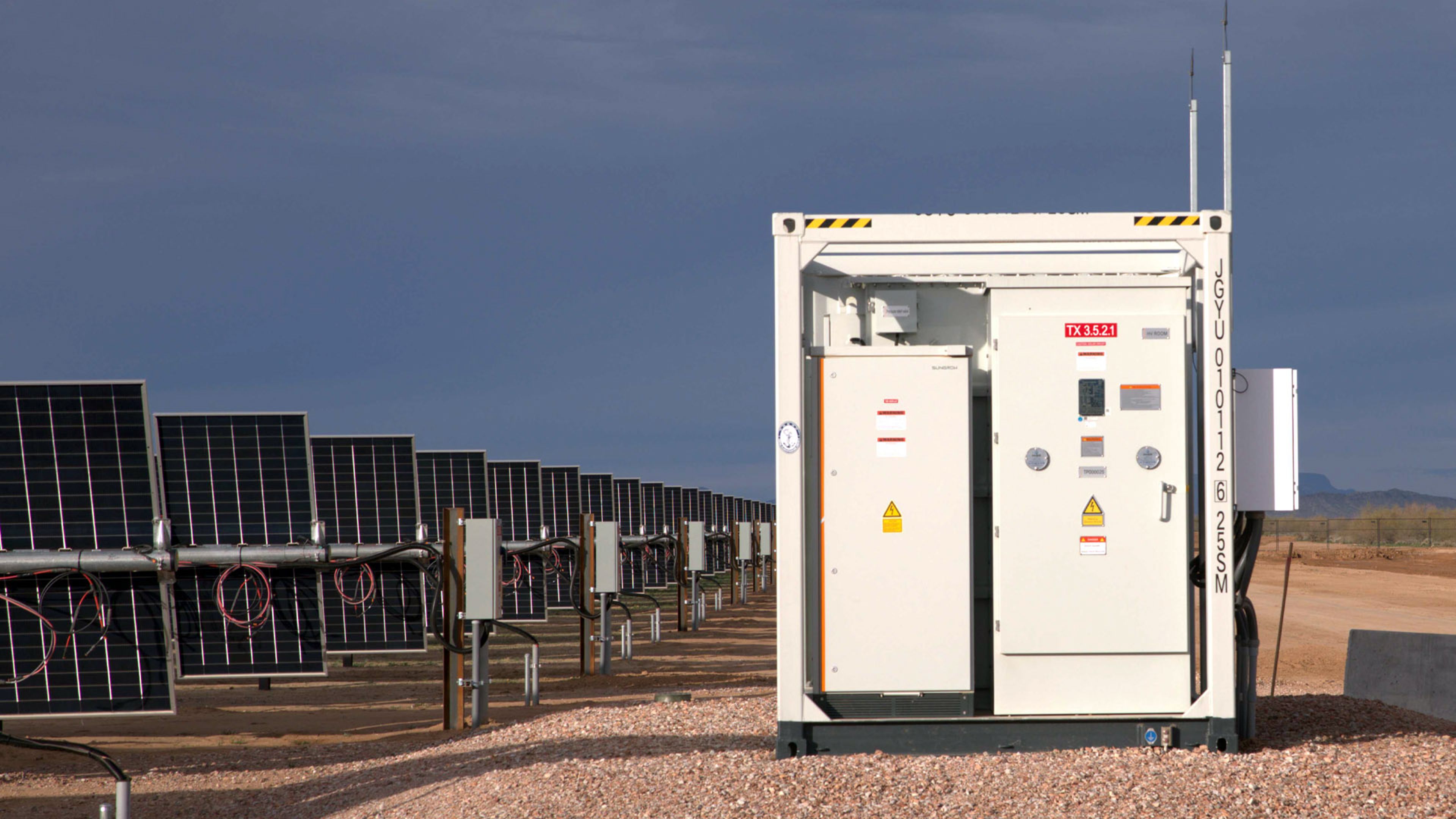 A row of solar panels in a desert landscape positioned beside a white electrical equipment container under a cloudy sky.