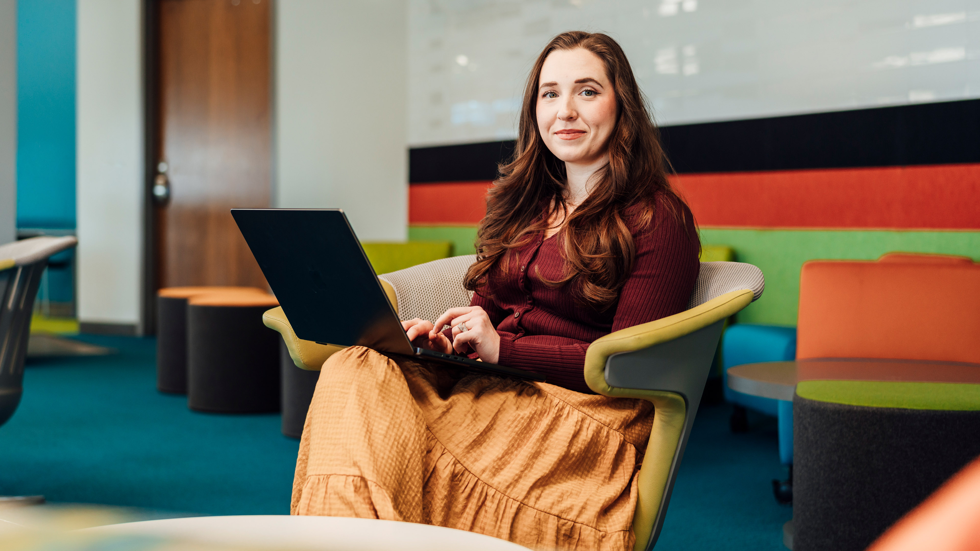 Person seated in a modern office lounge area holding a laptop, with colorful seating and workspace elements in the background