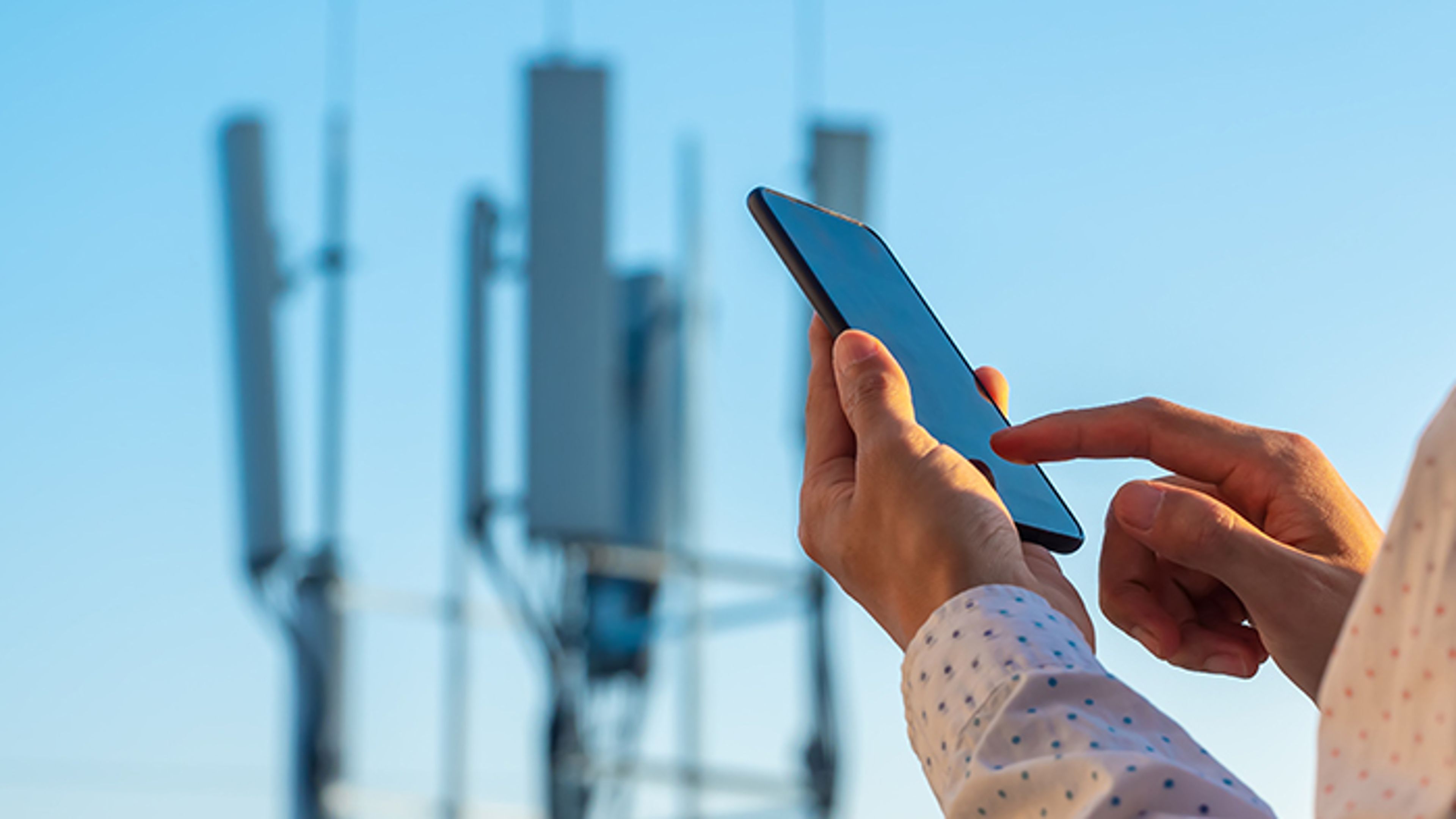 Hand with a mobile phone in the foreground, telecom tower blurred in the background