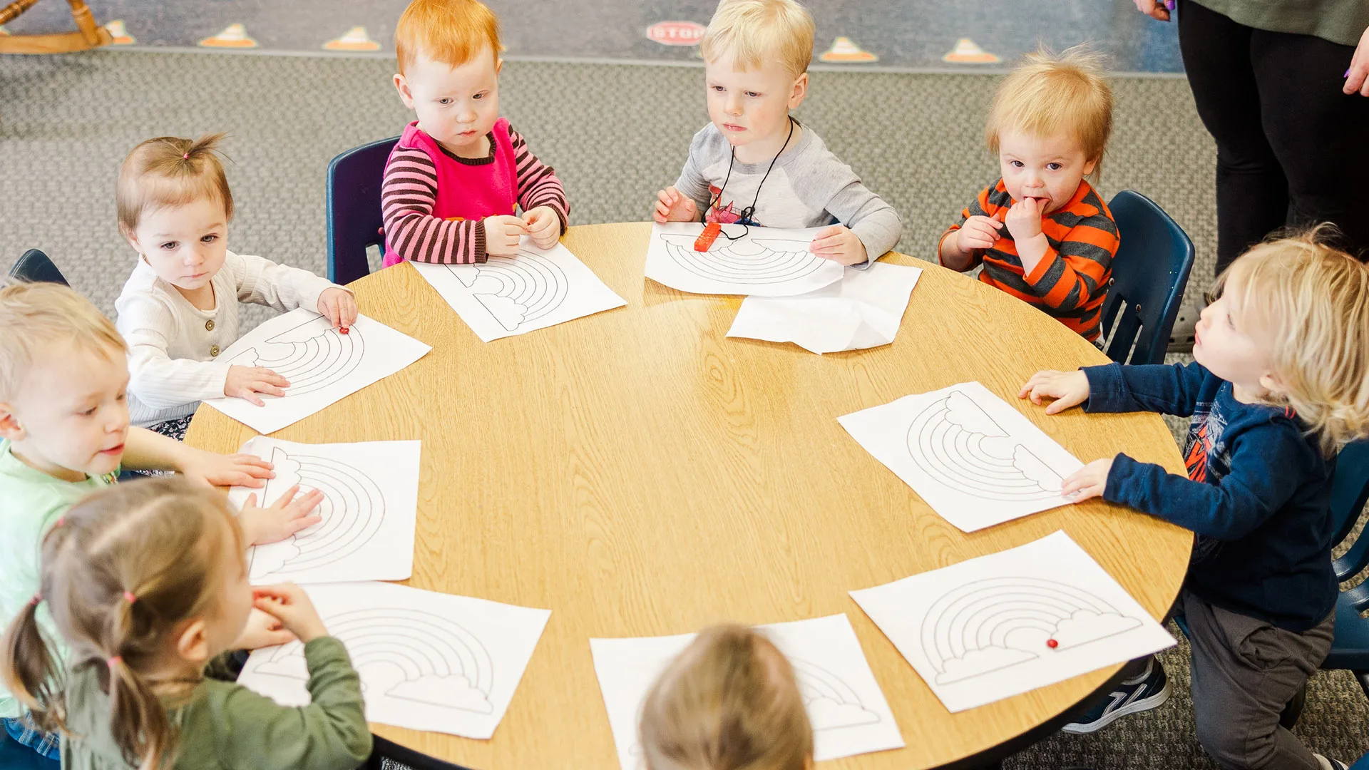 A group of young children sitting around a circular classroom table while coloring rainbow worksheets.