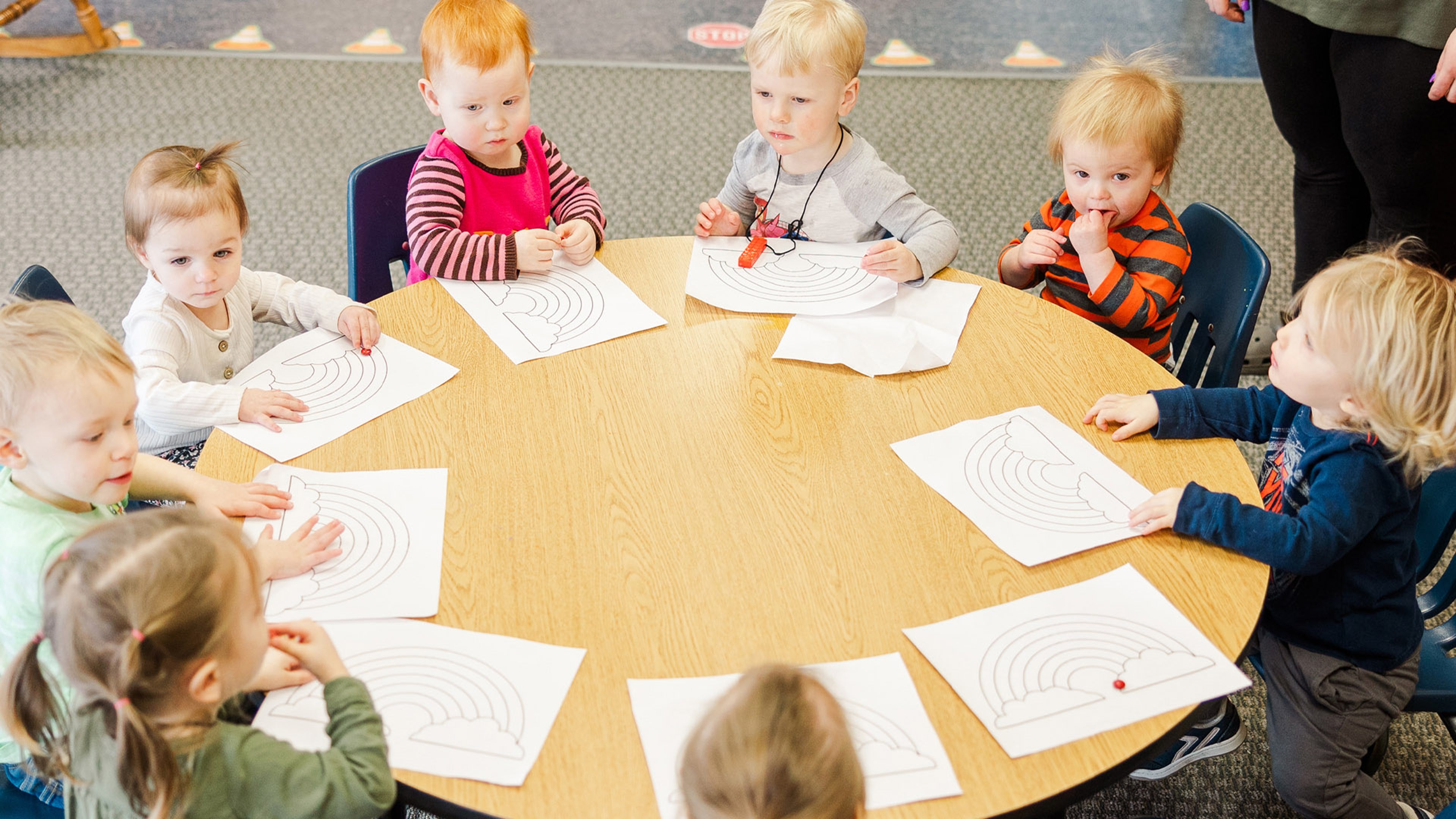 A group of young children sitting around a circular classroom table while coloring rainbow worksheets.
