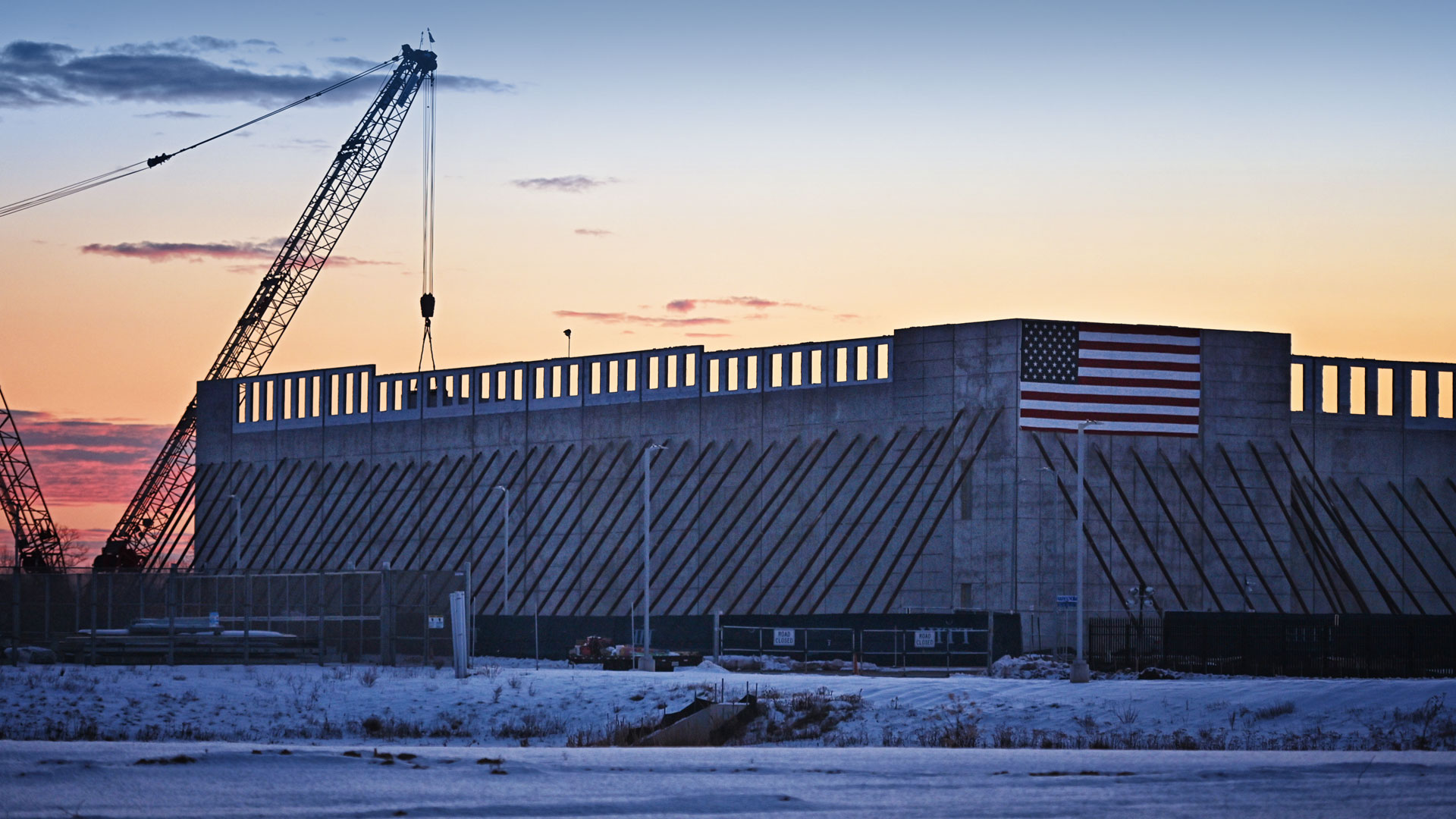 Data center facility under construction with a crane beside the building, set against a sunset sky, with an American flag mounted on the structure.