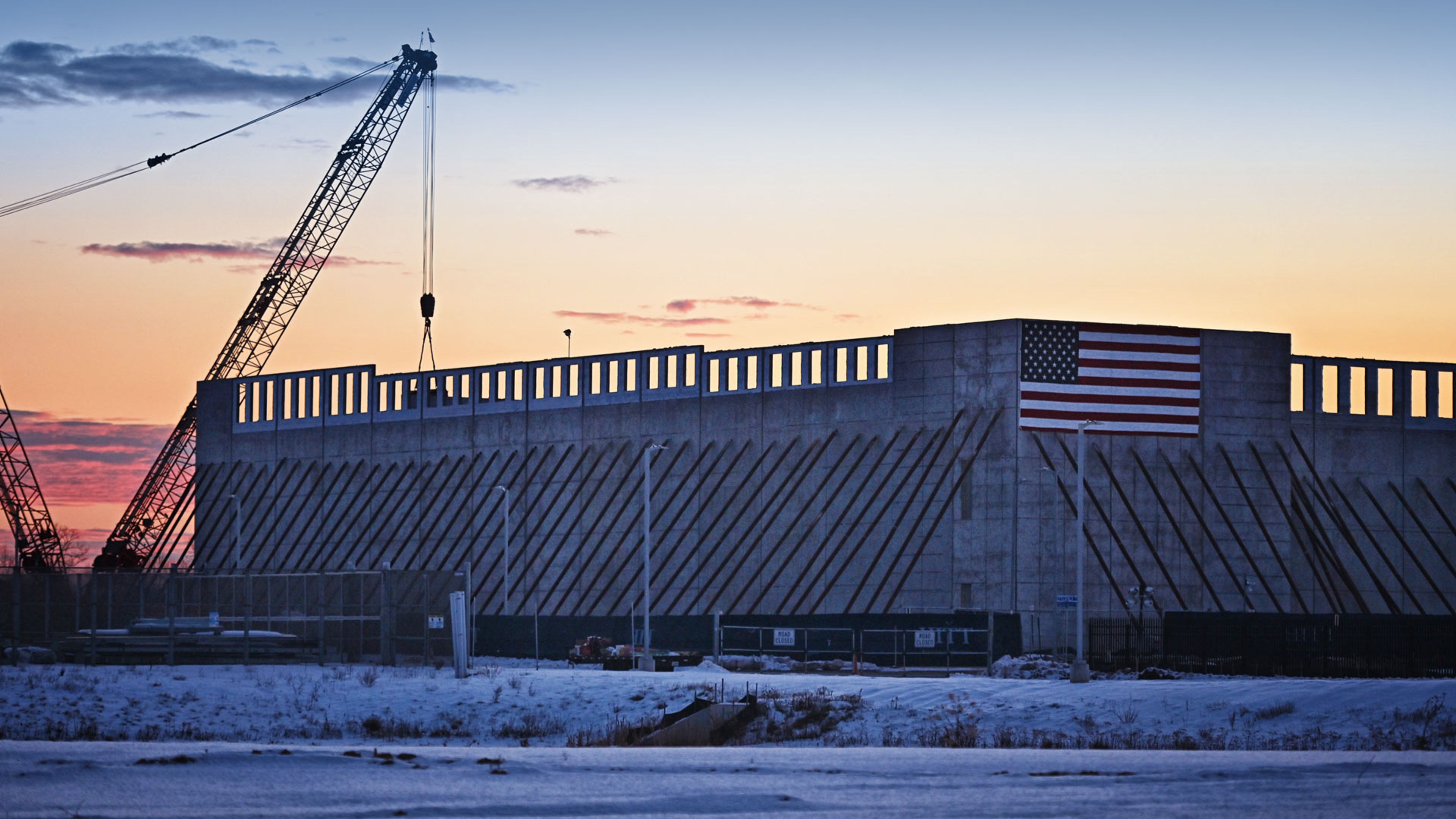 Data center facility under construction with a crane beside the building, set against a sunset sky, with an American flag mounted on the structure.