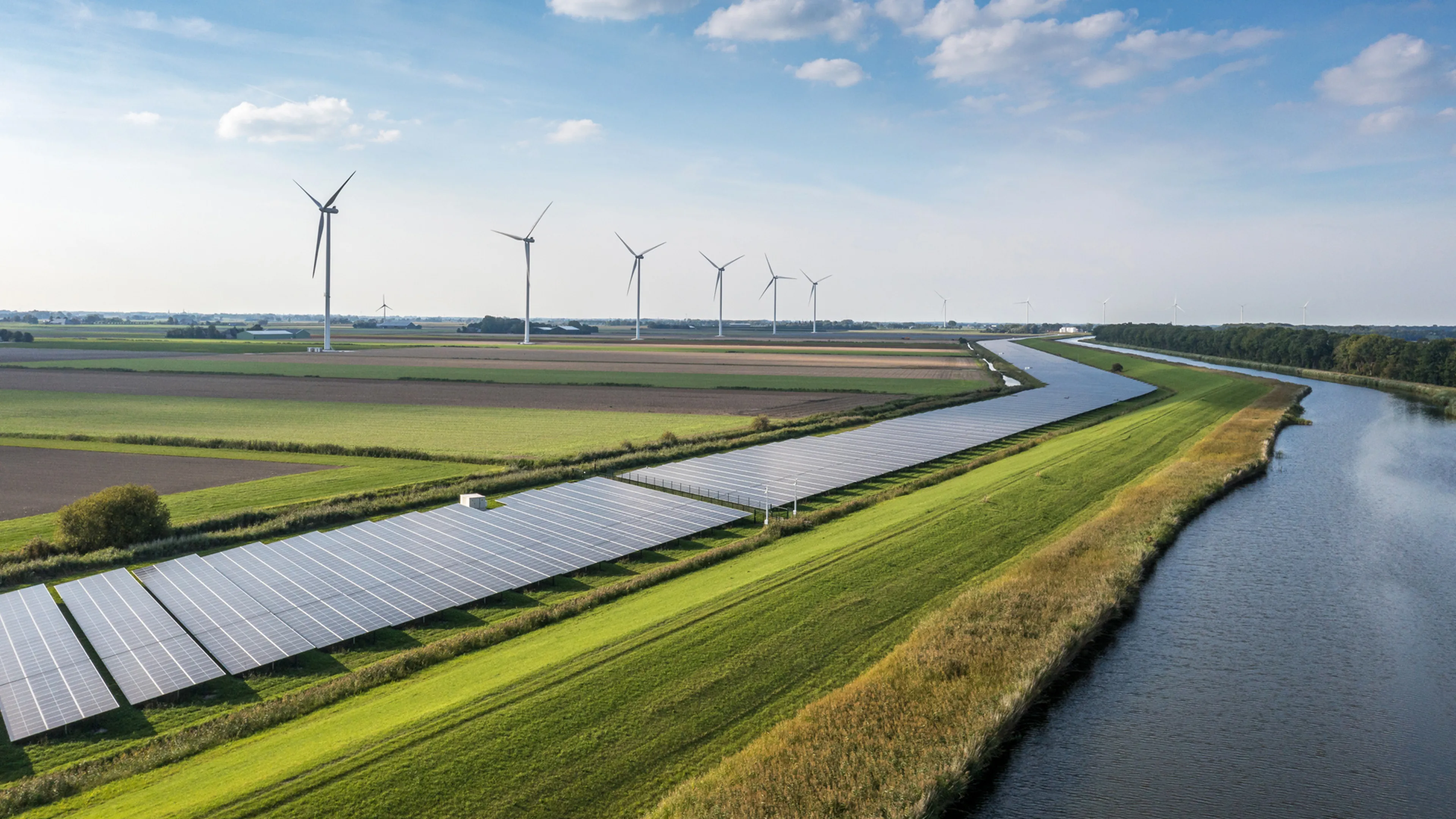 Wide landscape view of solar panels and wind turbines alongside a canal or waterway in an open rural setting