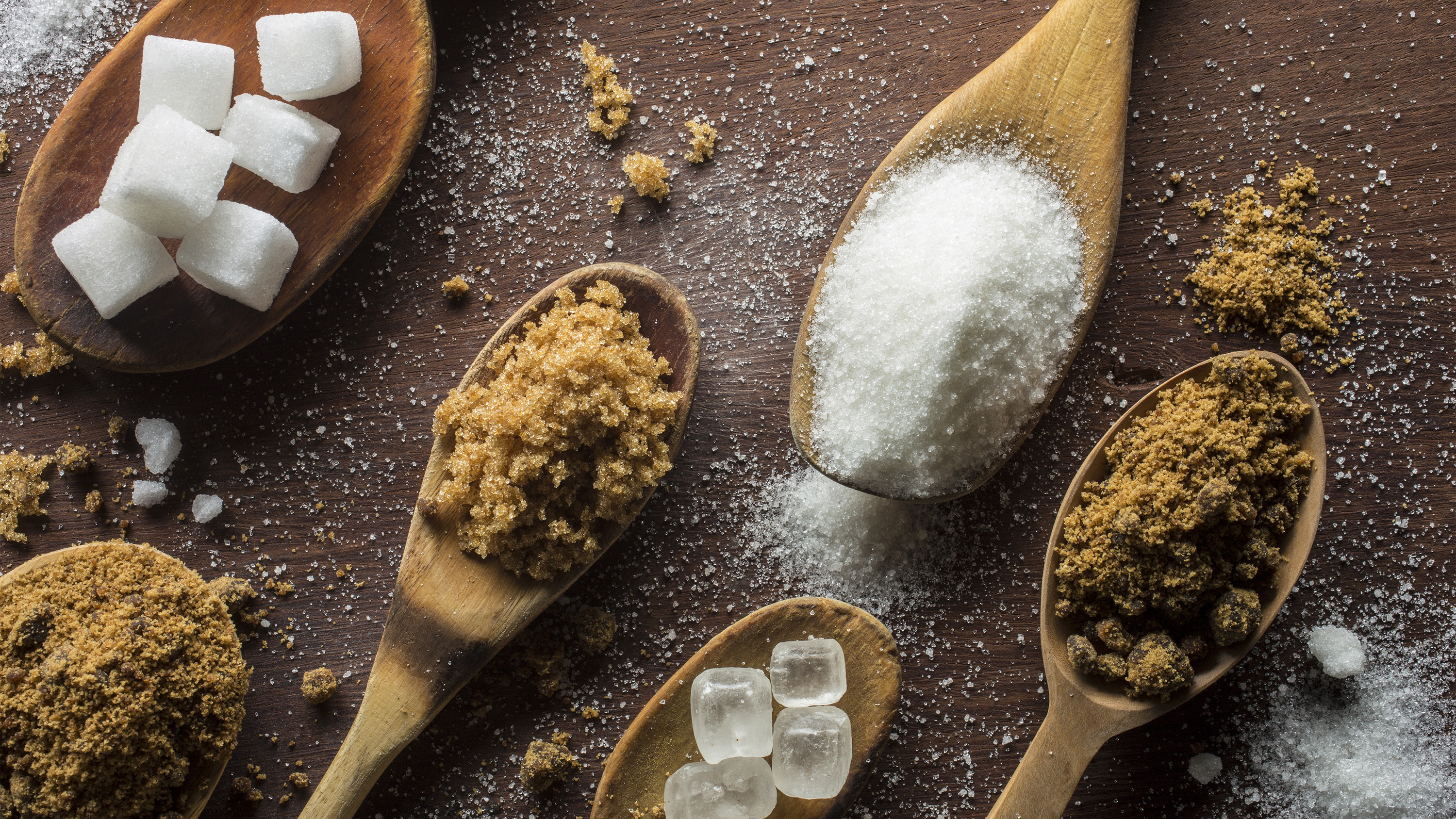 Six wooden spoons on table with different types of sugar, including sugar cubes, brown sugar, white granulated sugar, rock sugar and raw sugar
