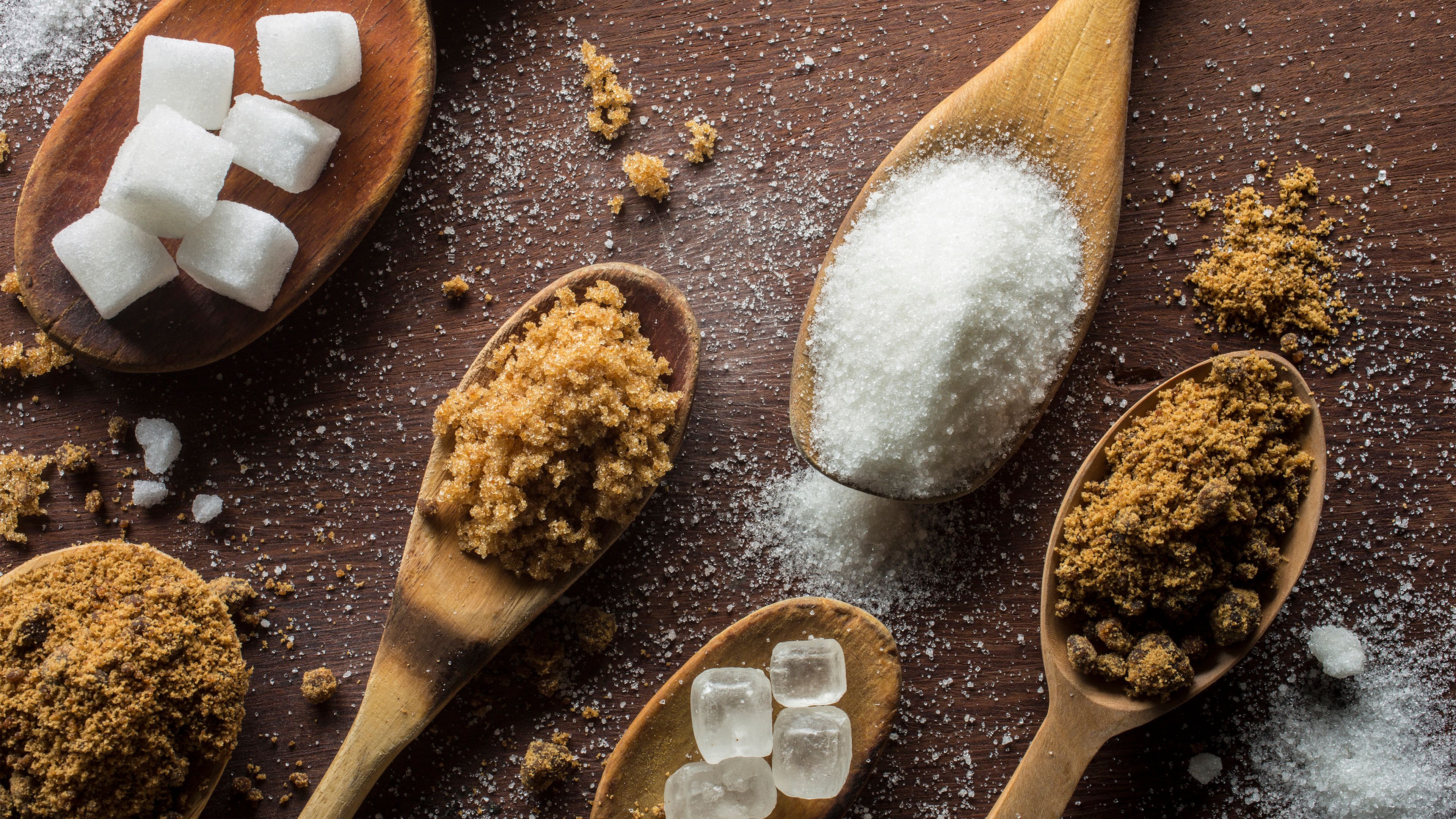 Six wooden spoons on table with different types of sugar, including sugar cubes, brown sugar, white granulated sugar, rock sugar and raw sugar