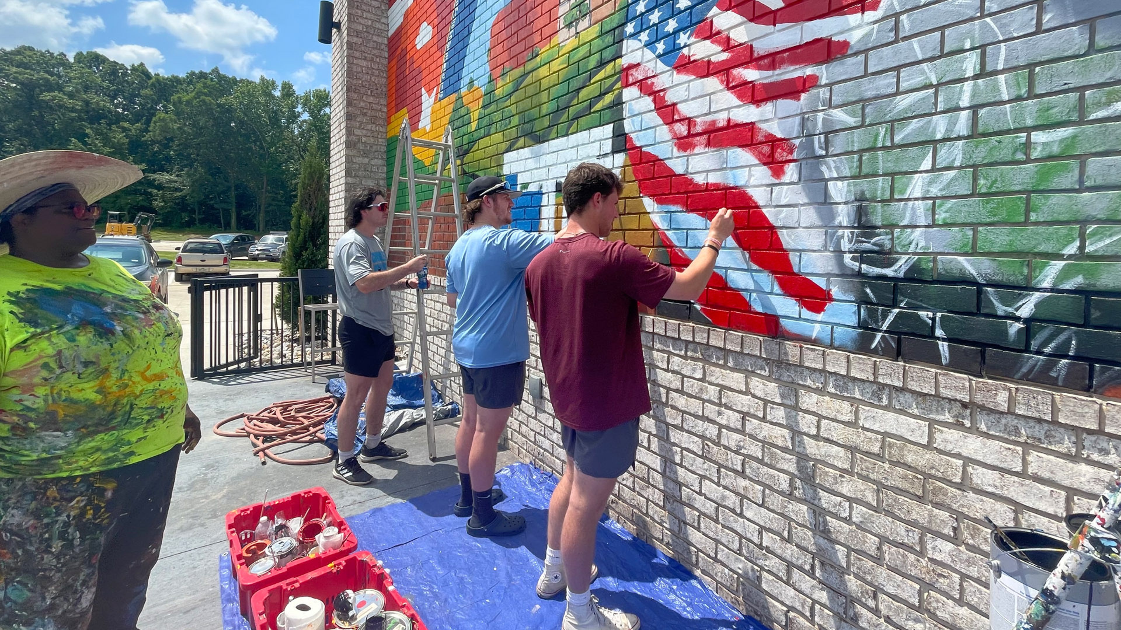 Several individuals painting a colorful outdoor mural on a brick wall using ladders and paint supplies.
